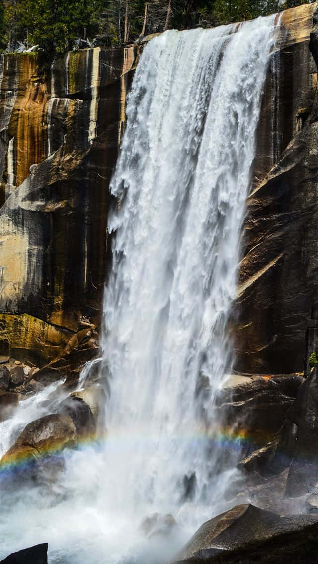 Valley 桌布 壁紙 — 一道強勁的瀑布從崎嶇的懸崖上傾瀉而下，在下方霧氣騰騰的水霧中形成一道鮮豔的彩虹。