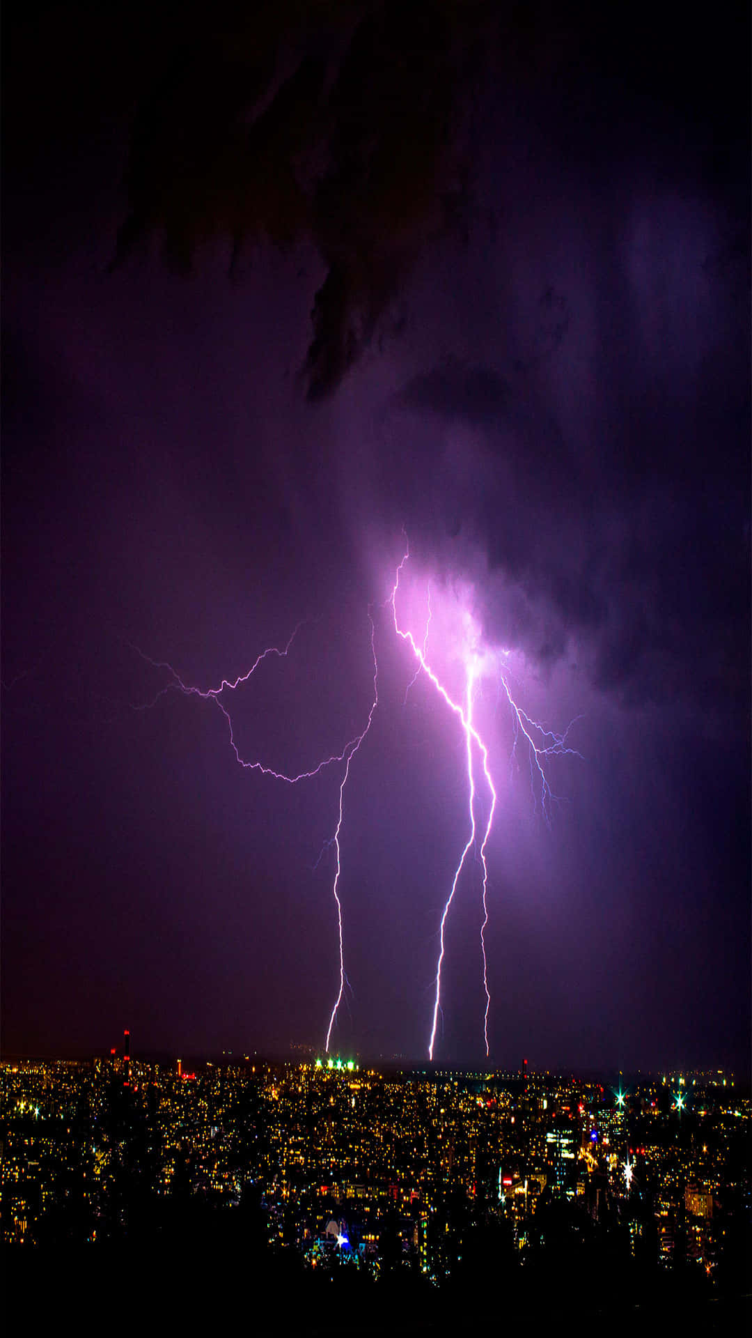 Purple Lightning 桌布 壁紙 — 一道強勁的紫色閃電照亮了暴風雨夜晚的天空，下方是燈火通明的廣闊城市景觀。
