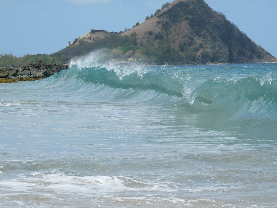 山景 桌布 壁紙 — 一道強勁的藍綠色海浪在岩石海岸附近翻捲並破碎，背景是崎嶇的山丘和晴朗的藍天。