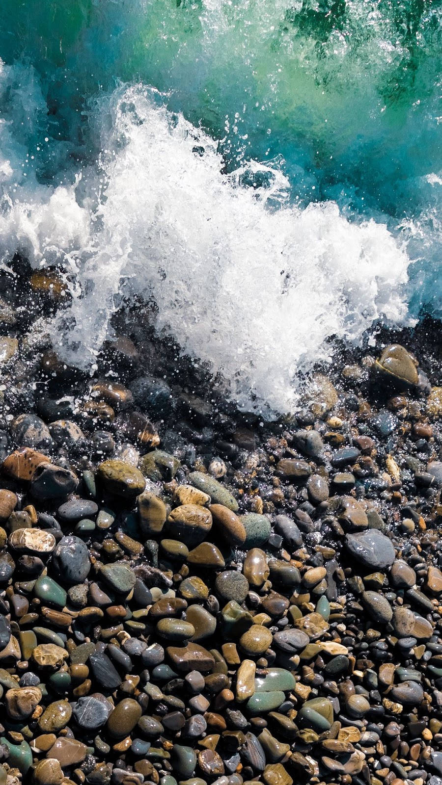 Wave 桌布 壁紙 — 一道強勁的藍綠色海浪拍打陽光照耀的鵝卵石海灘，白色泡沫在光滑的圓石上爆開。