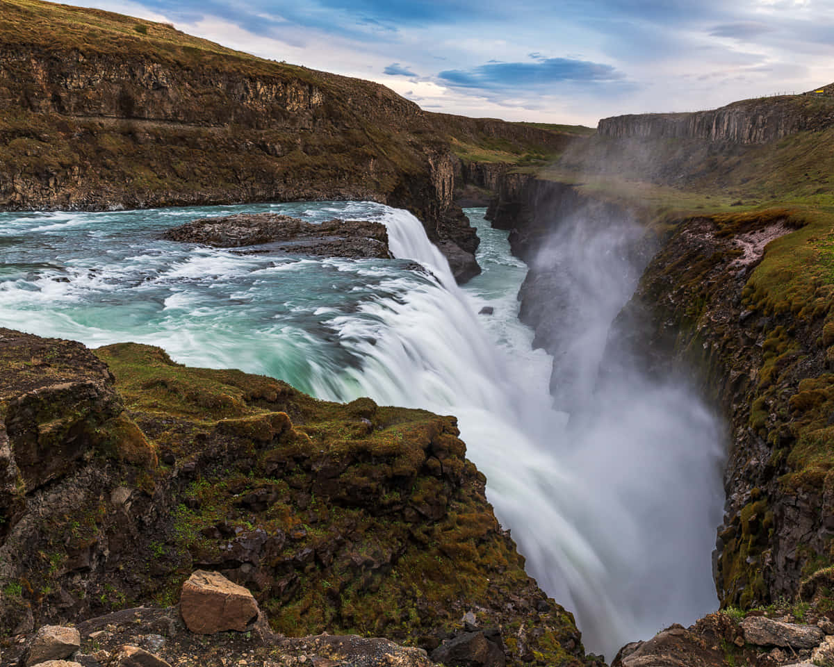 Iceland 桌布 壁紙 — 一道強勁的藍綠色瀑布衝入霧氣瀰漫的峽谷，兩旁是長滿苔蘚的懸崖，天空局部多雲。