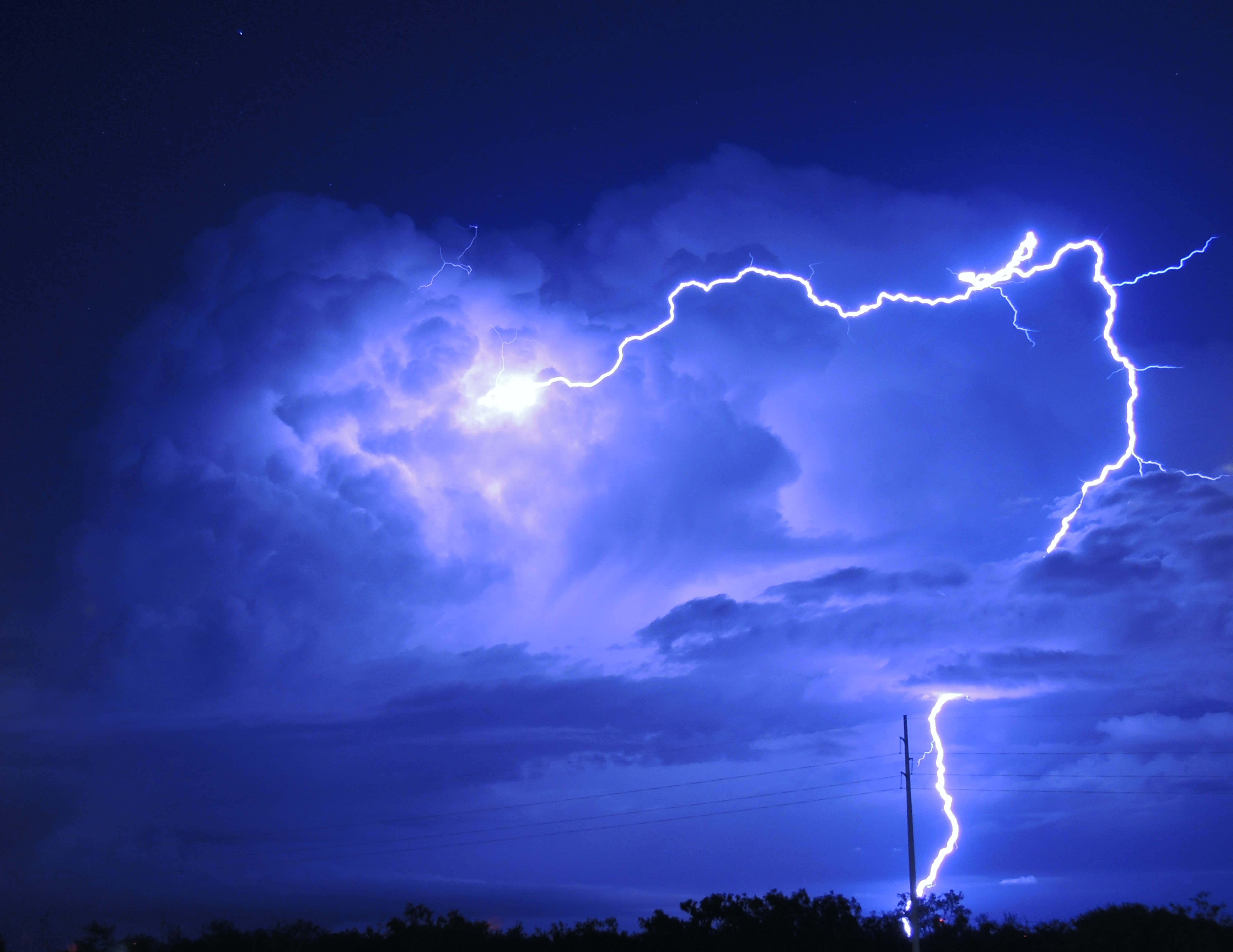 閃電 桌布 壁紙 — 一道強勁的閃電在黑暗的暴風雨天空中分叉，照亮了紫色的雲層和下方樹木的剪影。