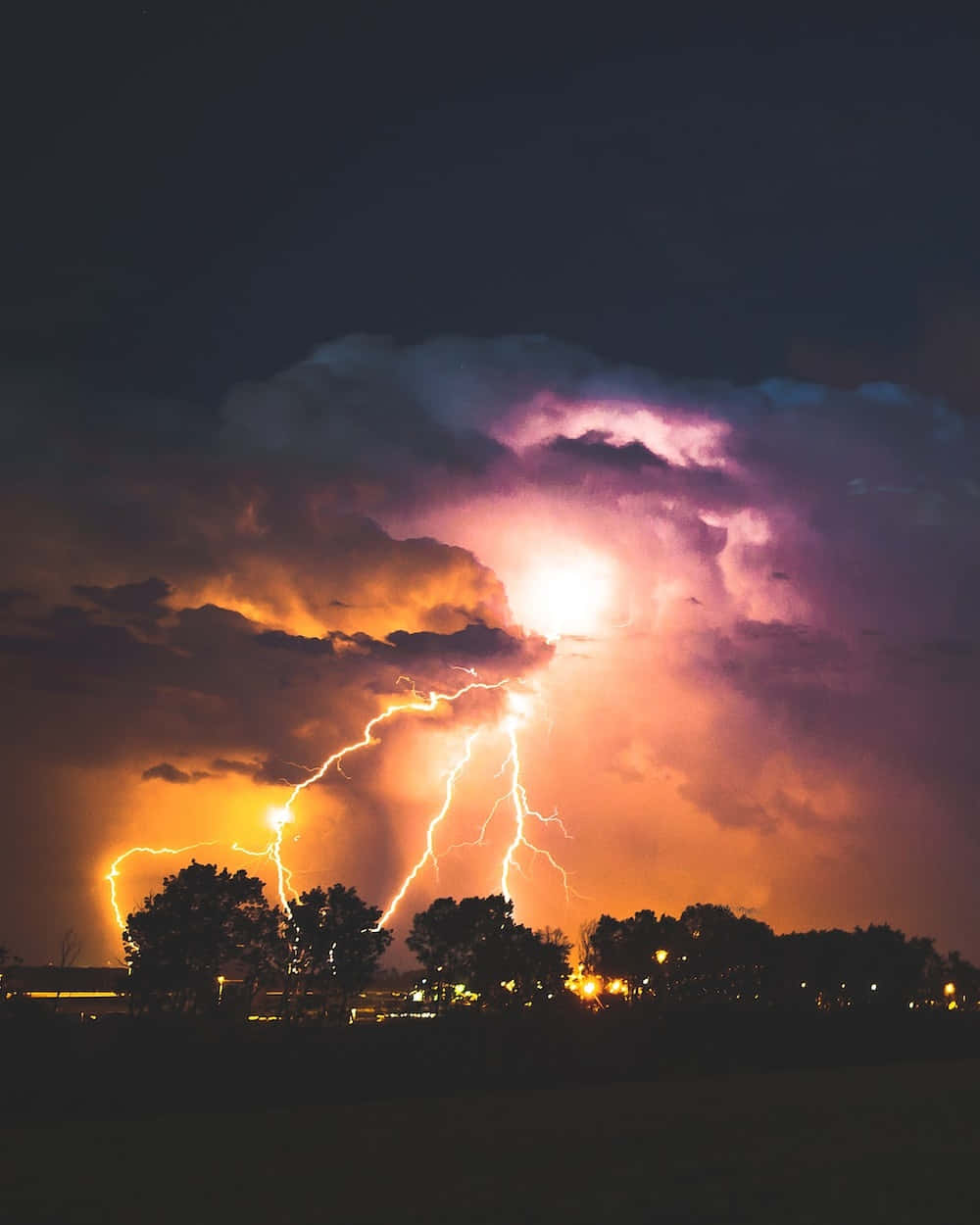 Lightning Bolt 桌布 壁紙 — 一道強勁的閃電照亮了暴風雨的夜空，紫色和橙色的雲層籠罩在剪影樹木和遠處燈光之上。