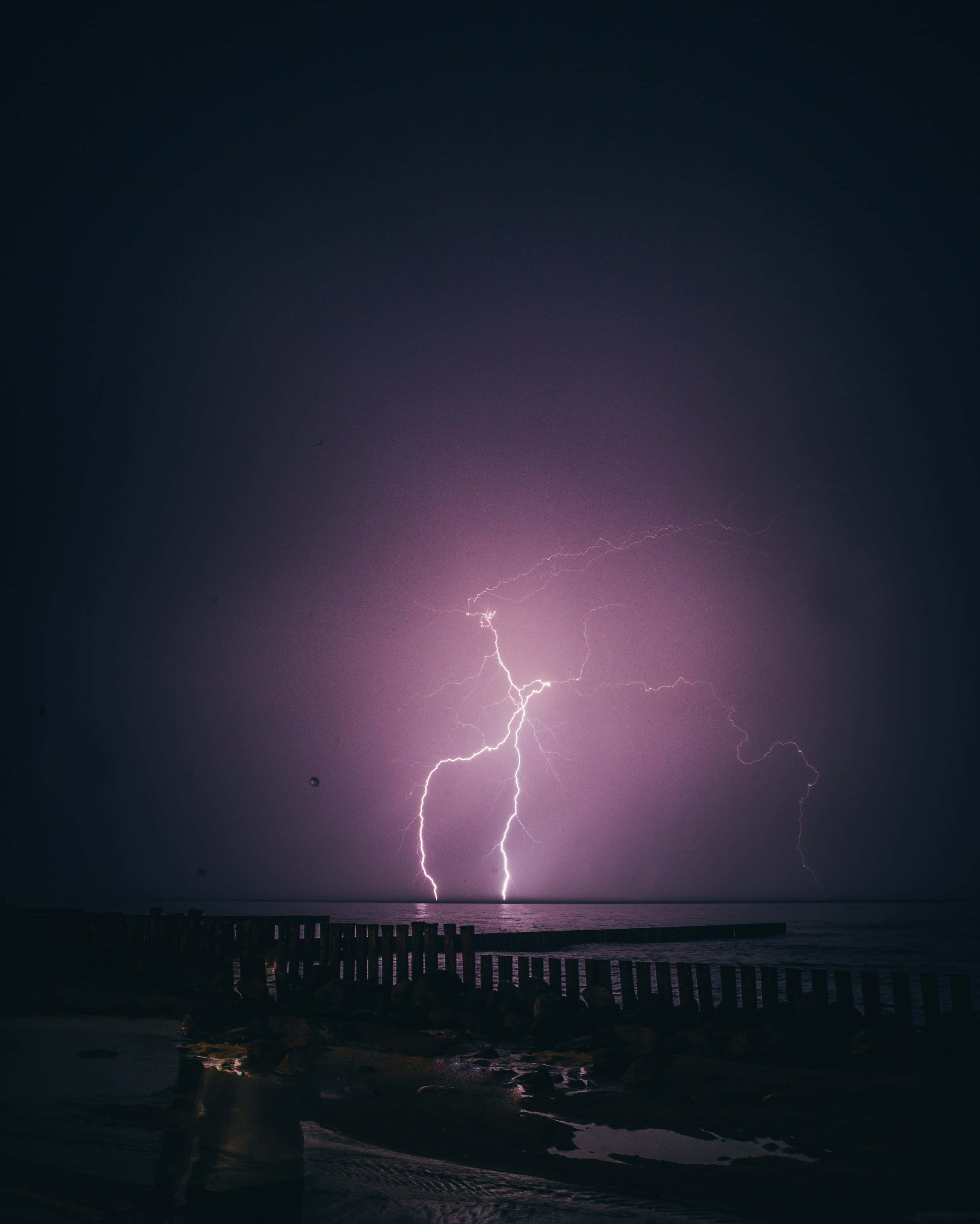 閃電 桌布 壁紙 — 一道強大的紫色閃電在夜間劃過黑暗的海洋，照亮了木製碼頭和濕漉漉的海岸線。