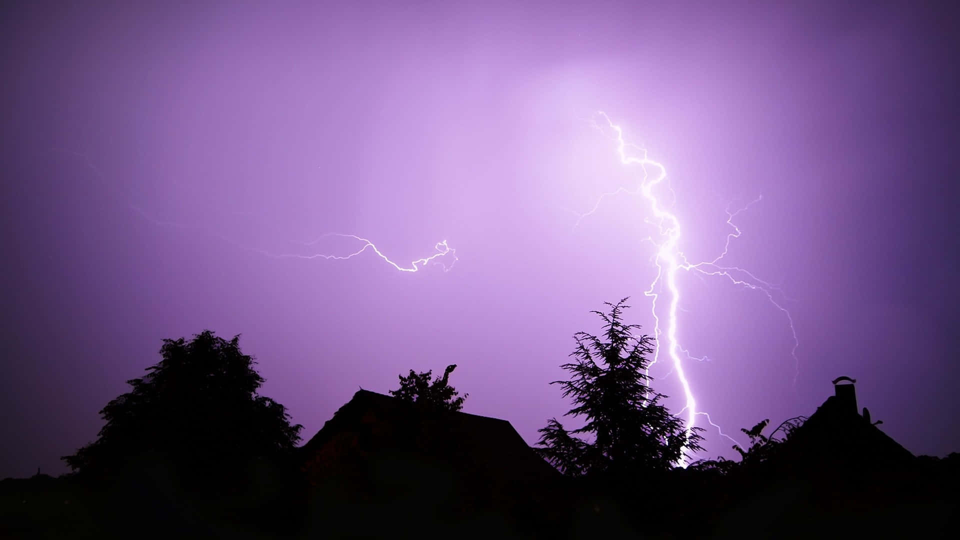 自然 桌布 壁紙 — 一道強大的閃電劃破紫羅蘭色的暴風雨天空，下方是剪影般的屋頂和樹木。
