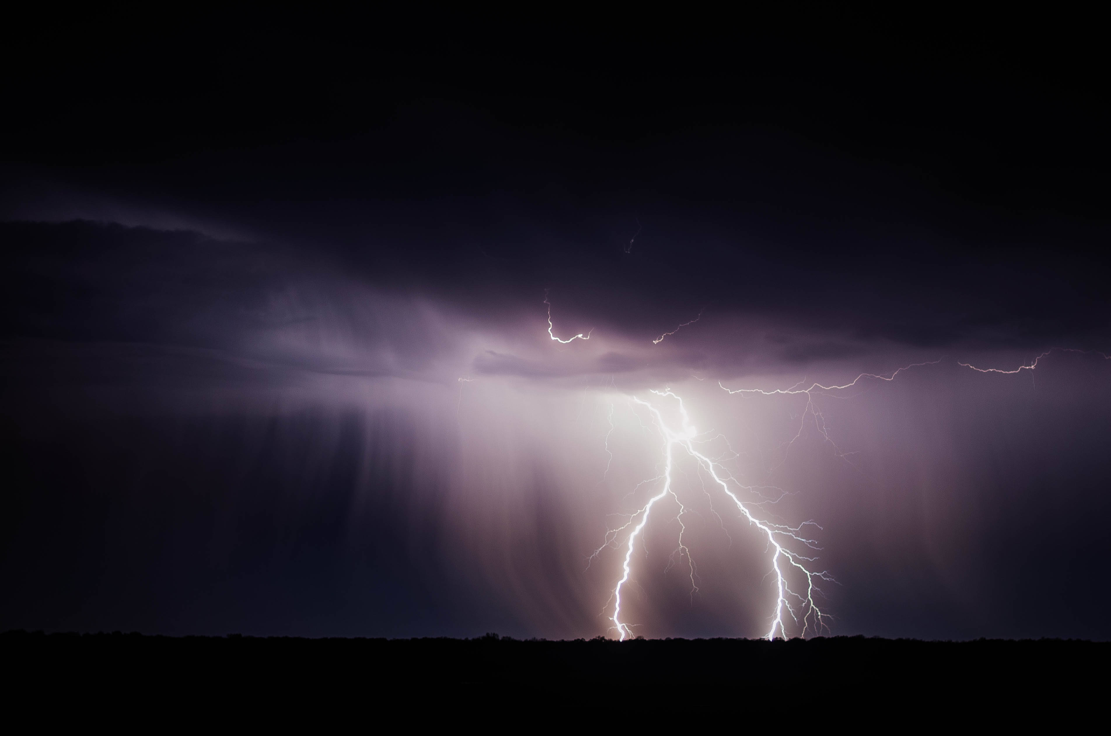 Storm 桌布 壁紙 — 一道強大的閃電劃破黑暗的暴風雨天空，映襯著剪影般的地平線，雨水傾瀉而下。