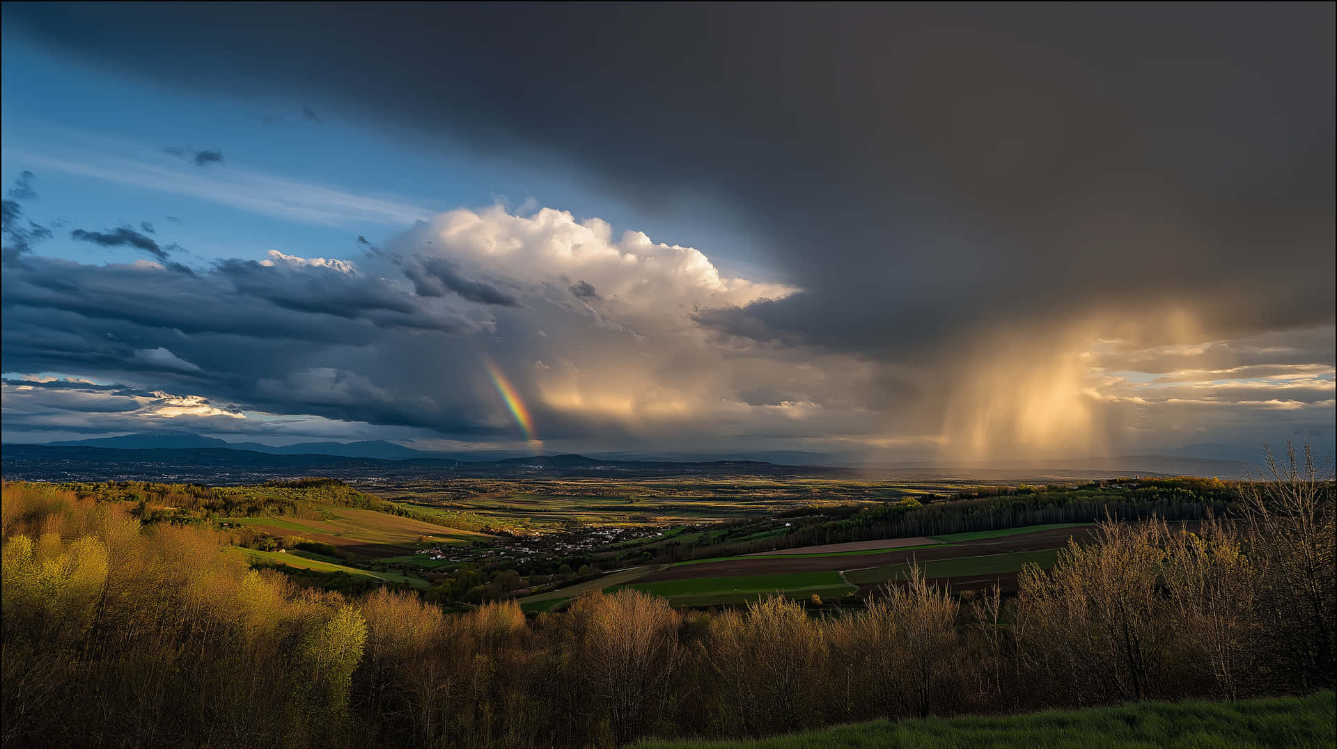 Dramatic Stormy Sky with Rainbow Over Green Landscape 壁紙 — 一道戲劇性的彩虹穿過暴風雲，拱形在陽光照耀的山谷上，有起伏的田野和遠山。