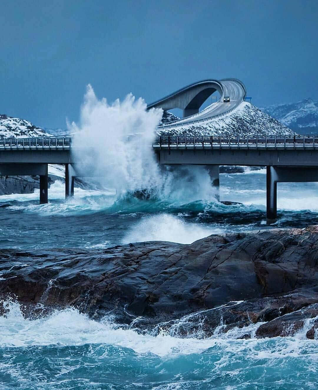 Road 桌布 壁紙 — 一道戲劇性的海浪拍打著崎嶇的岩石，背景是雪覆蓋的山脈和一座現代化的拱橋。