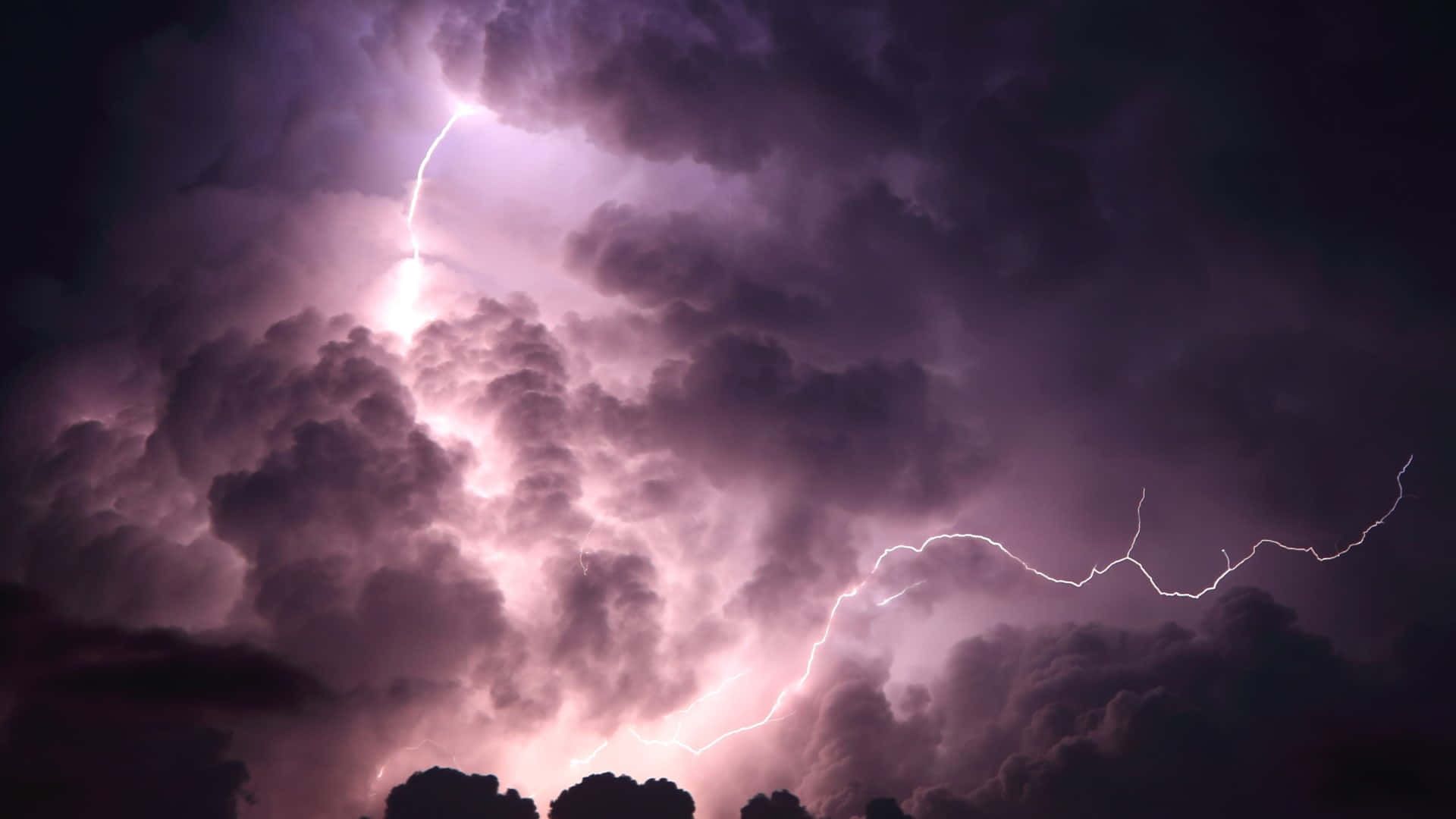 雲朵 桌布 壁紙 — 一道戲劇性的紫色閃電劈開高聳的暴風雲，下方是夜晚的黑暗樹線。