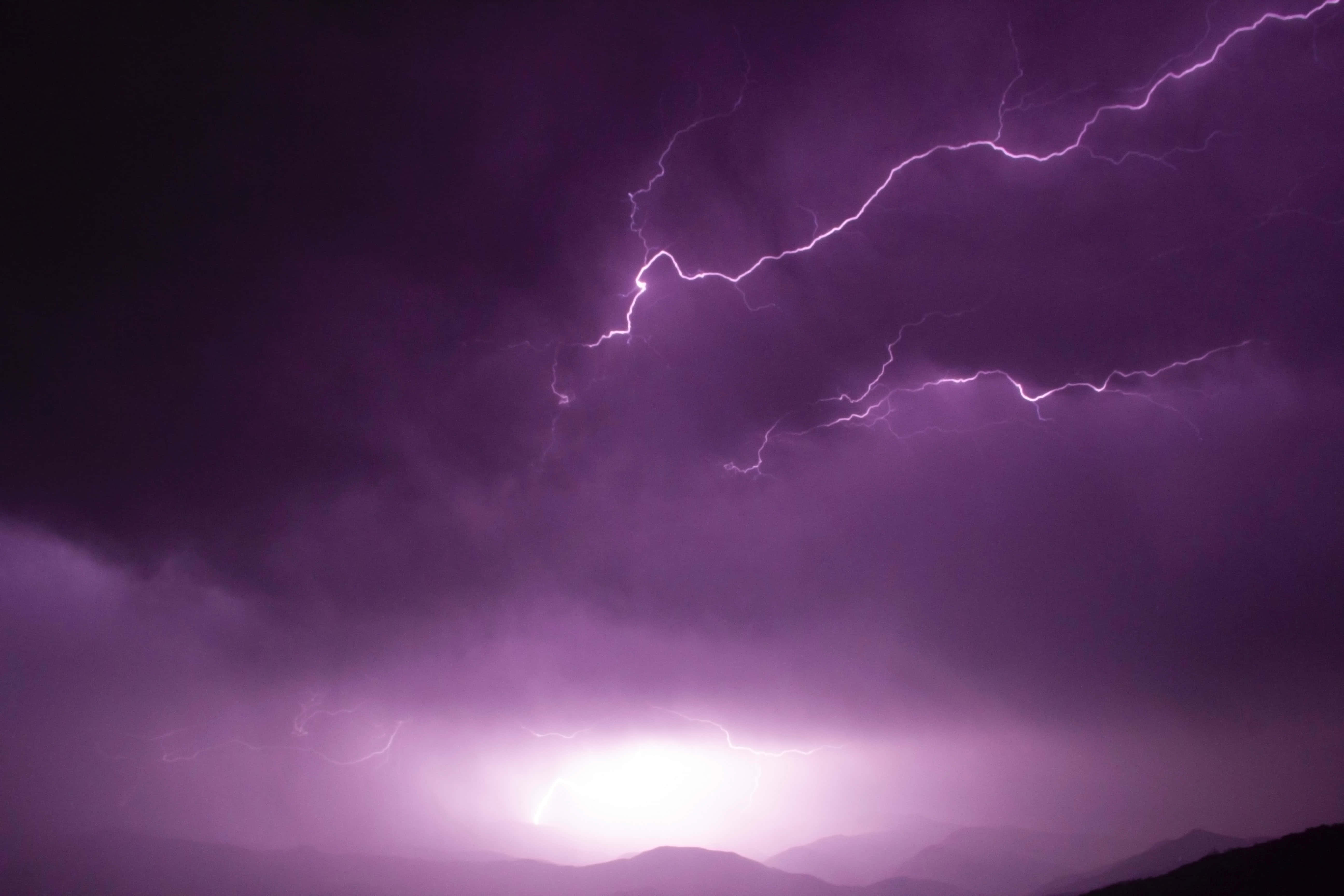 暗紫色美學 桌布 壁紙 — 一道戲劇性的紫色閃電在夜晚的暴風雨天空中，在剪影般的山脈上方分叉。