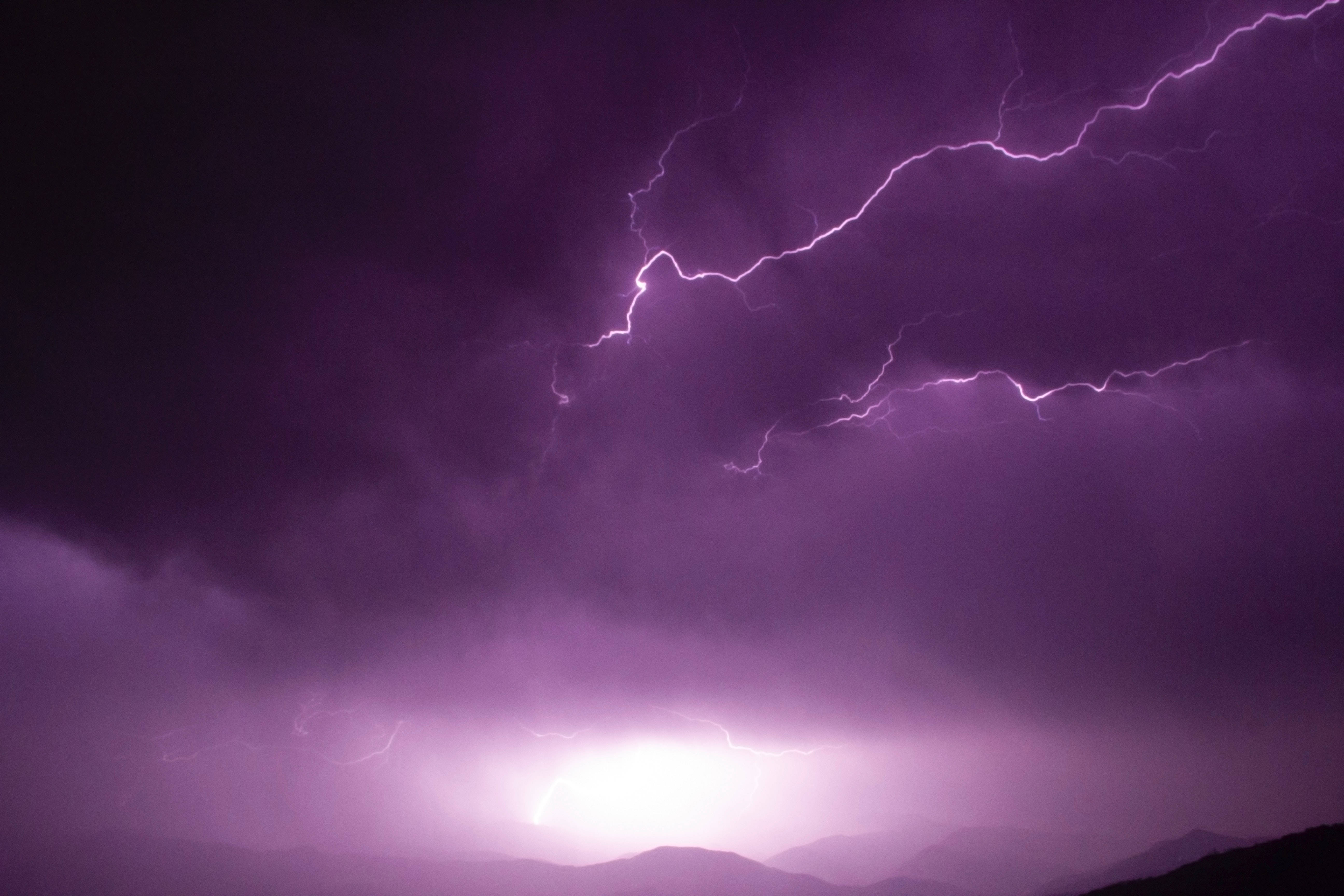 霓虹紫 iPhone 桌布 壁紙 — 一道戲劇性的紫色閃電在夜晚的暴風雨天空中，於剪影般的山脈上方分叉。