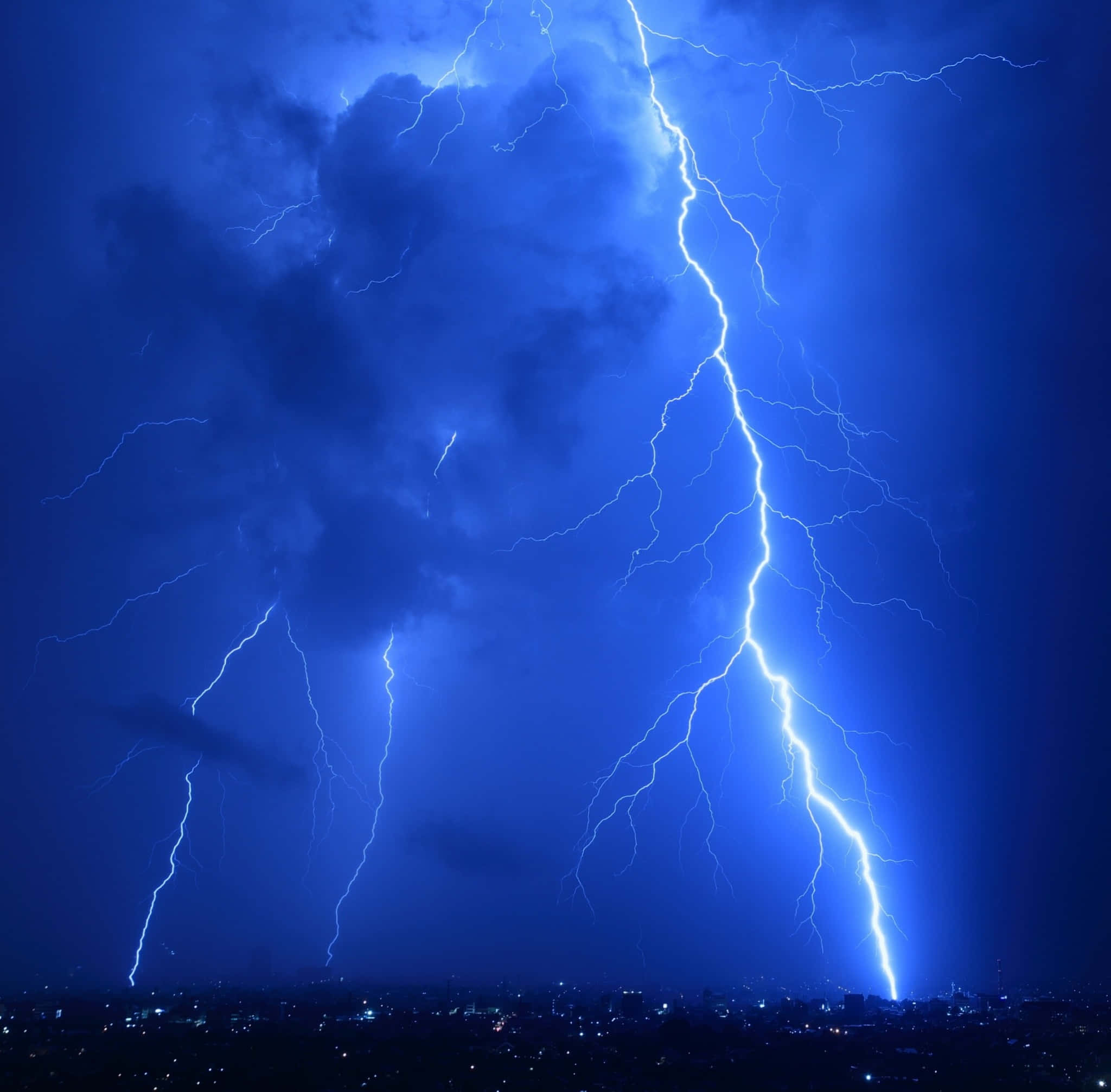 Coolest 桌布 壁紙 — 一道戲劇性的閃電在暴風雨的夜空中分叉，照亮了遠方燈火輝煌的城市天際線。