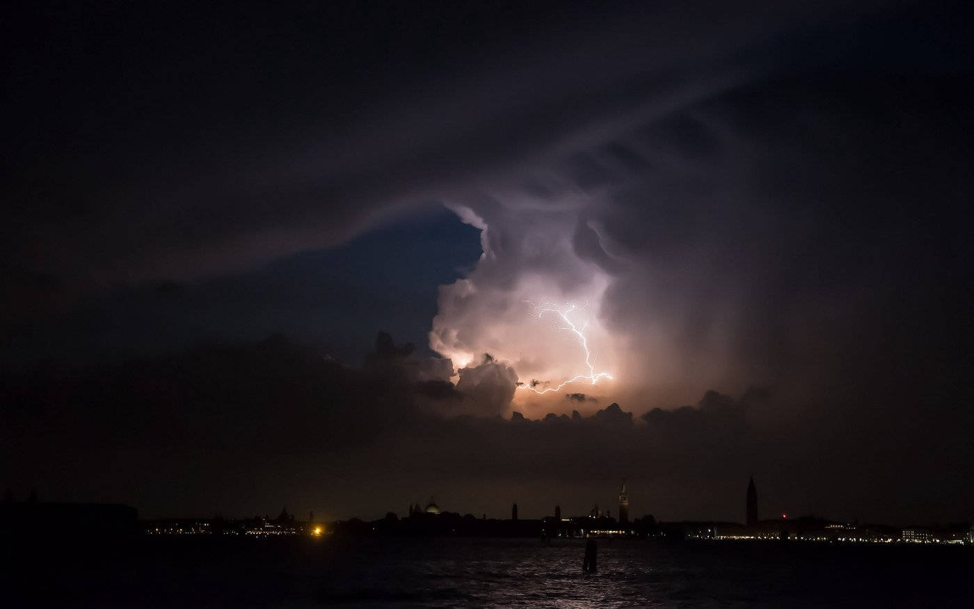 Storm 桌布 壁紙 — 一道戲劇性的閃電照亮了夜間城市天際線上方的黑暗暴風雲，水邊閃爍著燈光。
