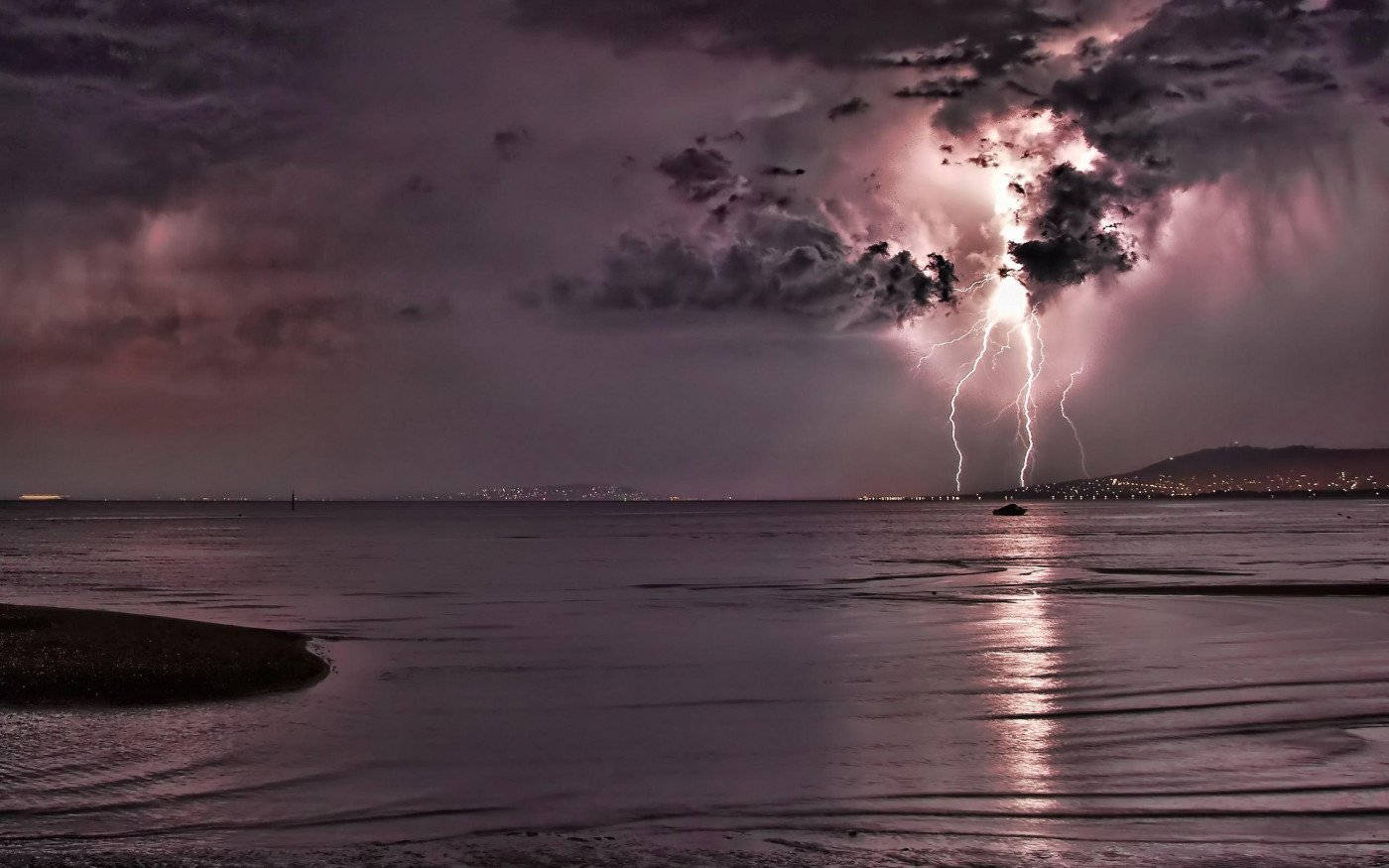 閃電 桌布 壁紙 — 一道戲劇性的閃電照亮了平靜海面上的暴風雨之夜，遠方地平線上有城市燈光。