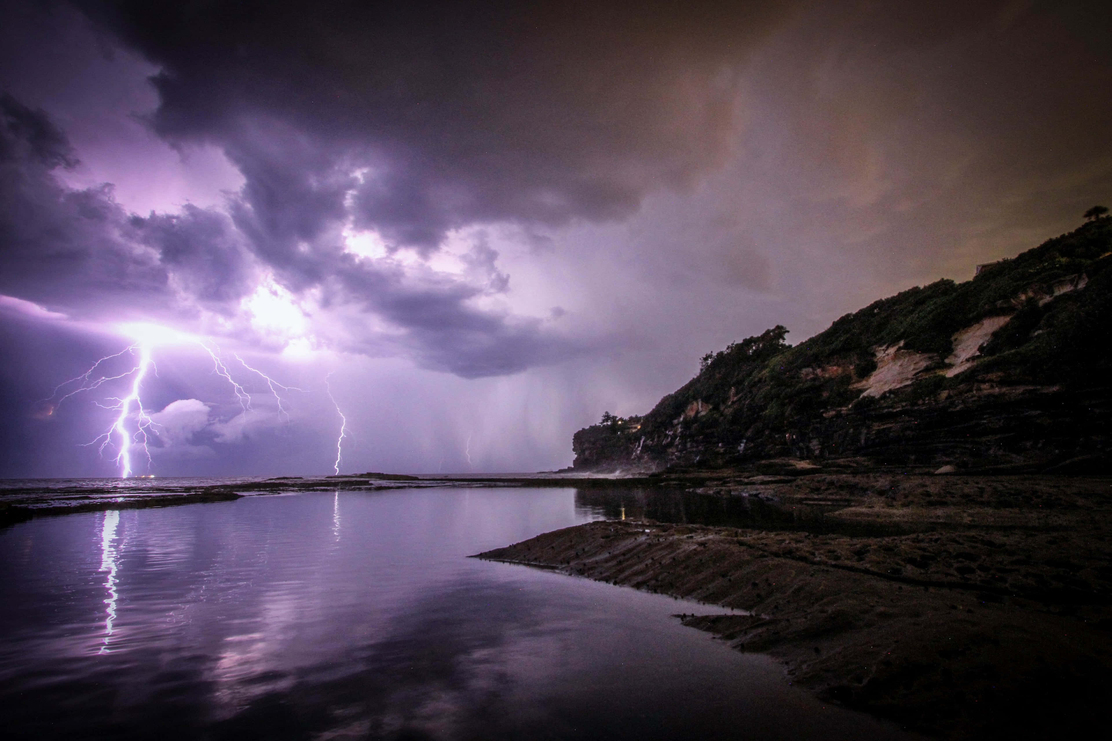 閃電 桌布 壁紙 — 一道戲劇性的閃電照亮了暴風雨之夜，平靜的海灣上有岩石峭壁。