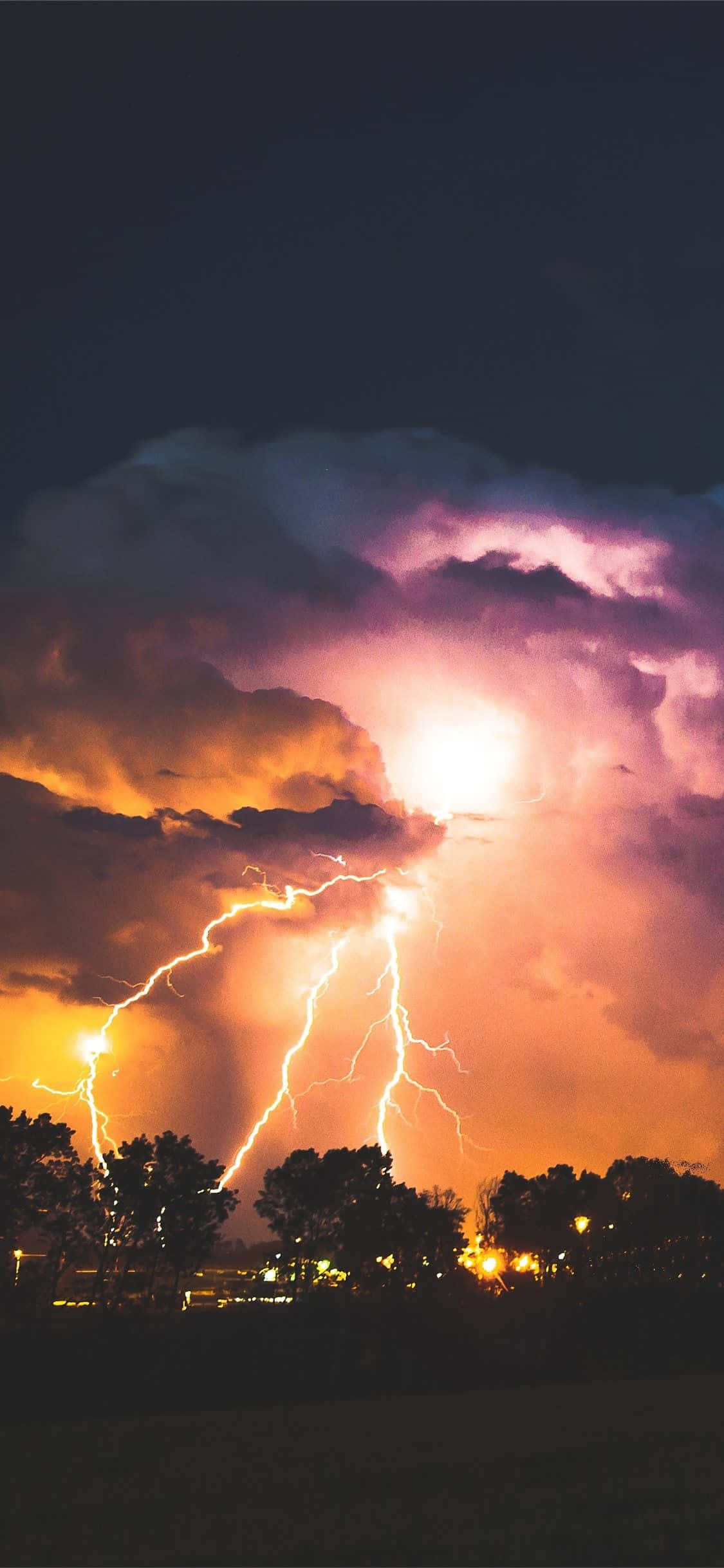 Lightning Bolt iPhone 桌布 壁紙 — 一道戲劇性的閃電照亮了暴風雨的夜空，紫色和橙色的雲層籠罩著剪影樹木和遠處的燈光。