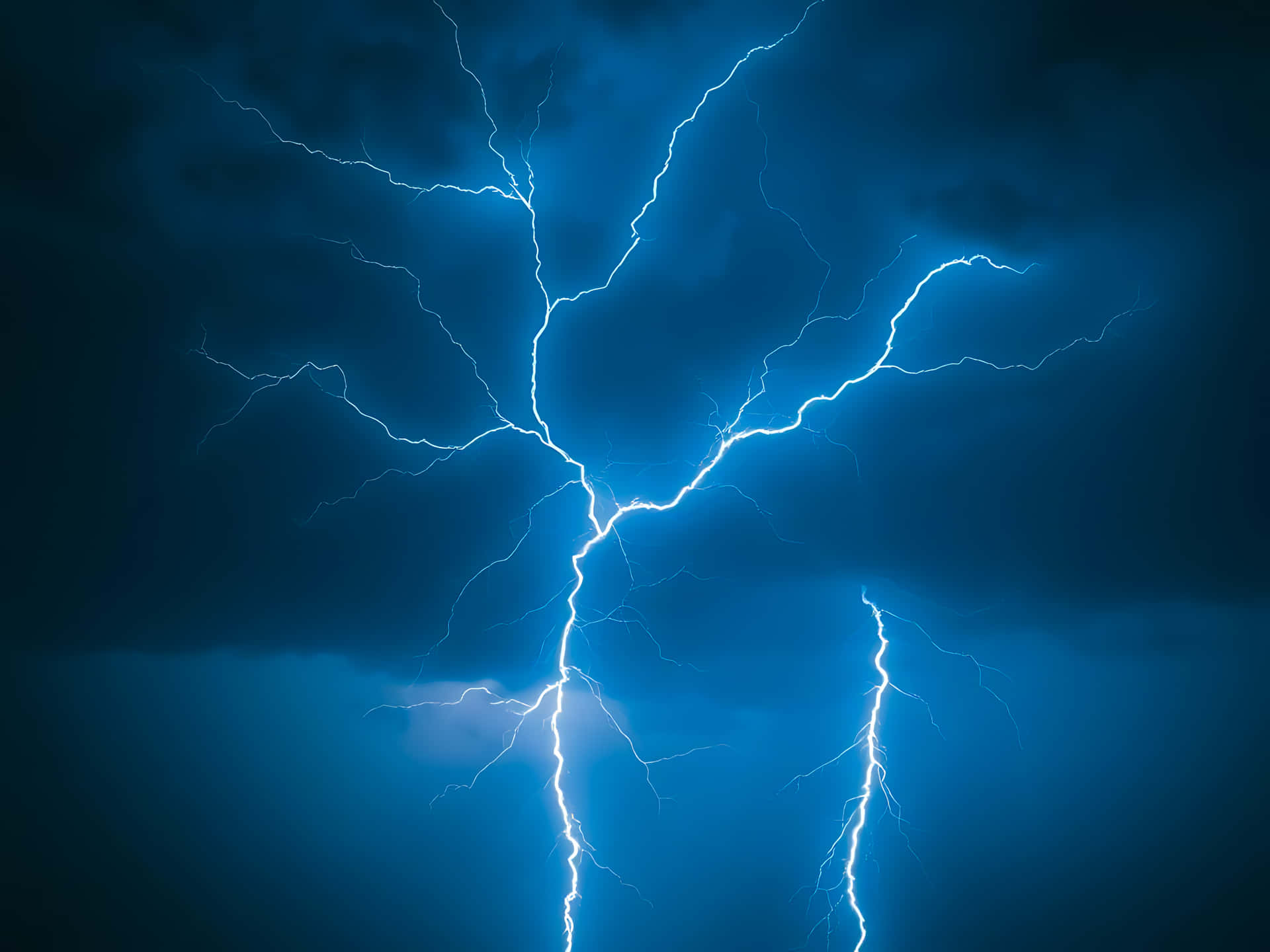 Lightning strikes illuminating the dark cloudy sky with electric power 壁紙 — 一道明亮的白色閃電在深藍色的暴風雨天空中分叉，帶有鋸齒狀的電枝。
