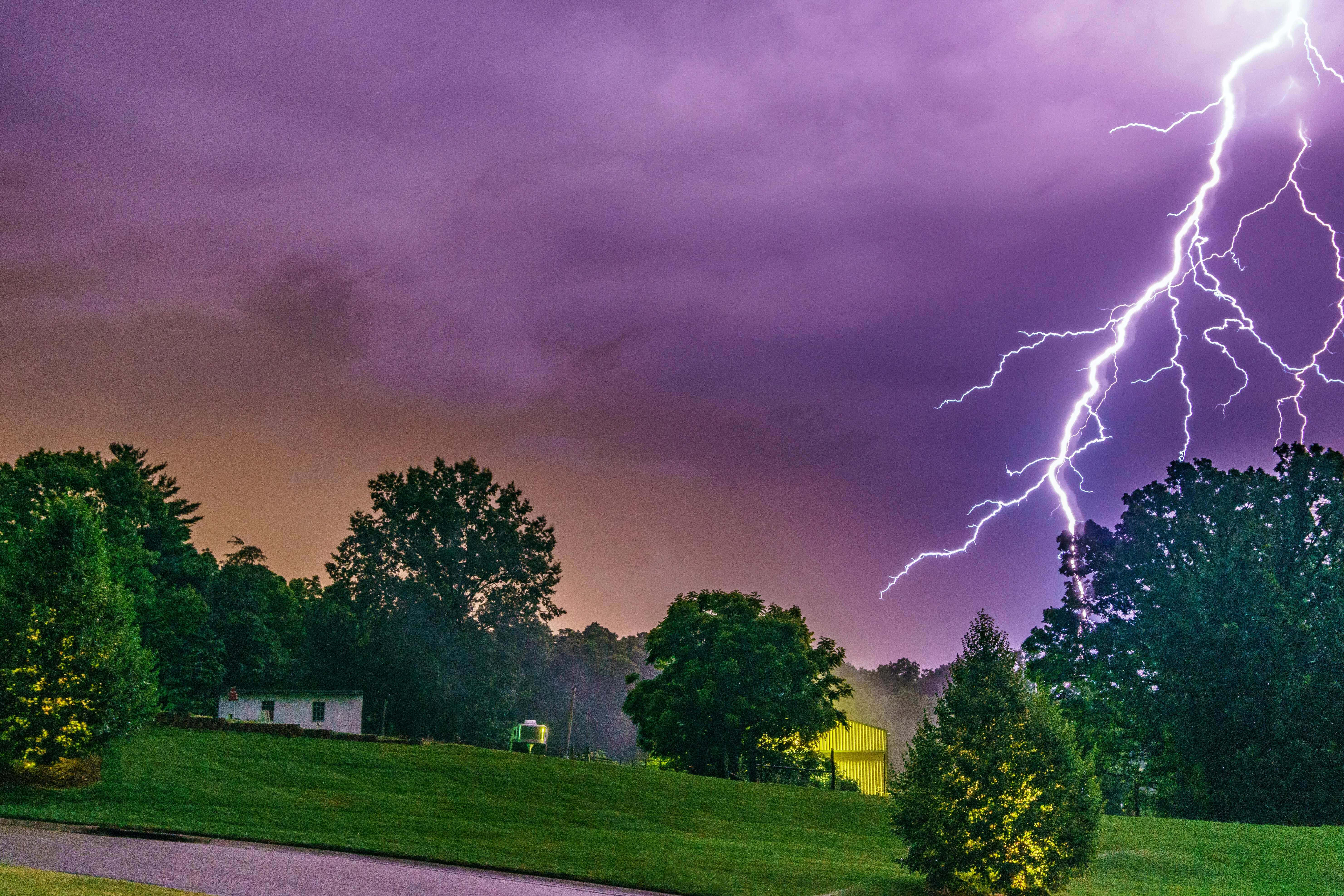 Lightning Bolt 桌布 壁紙 — 一道明亮的閃電劃破紫色暴風雨的天空，下方是寧靜的郊區景觀，有樹木和房屋。