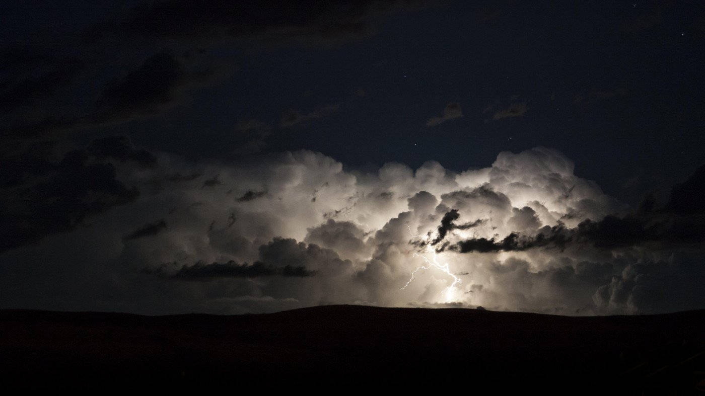  壁紙 — 一道明亮的閃電劈開巨大的暴風雲，籠罩在星空下黑暗的山丘輪廓之上。