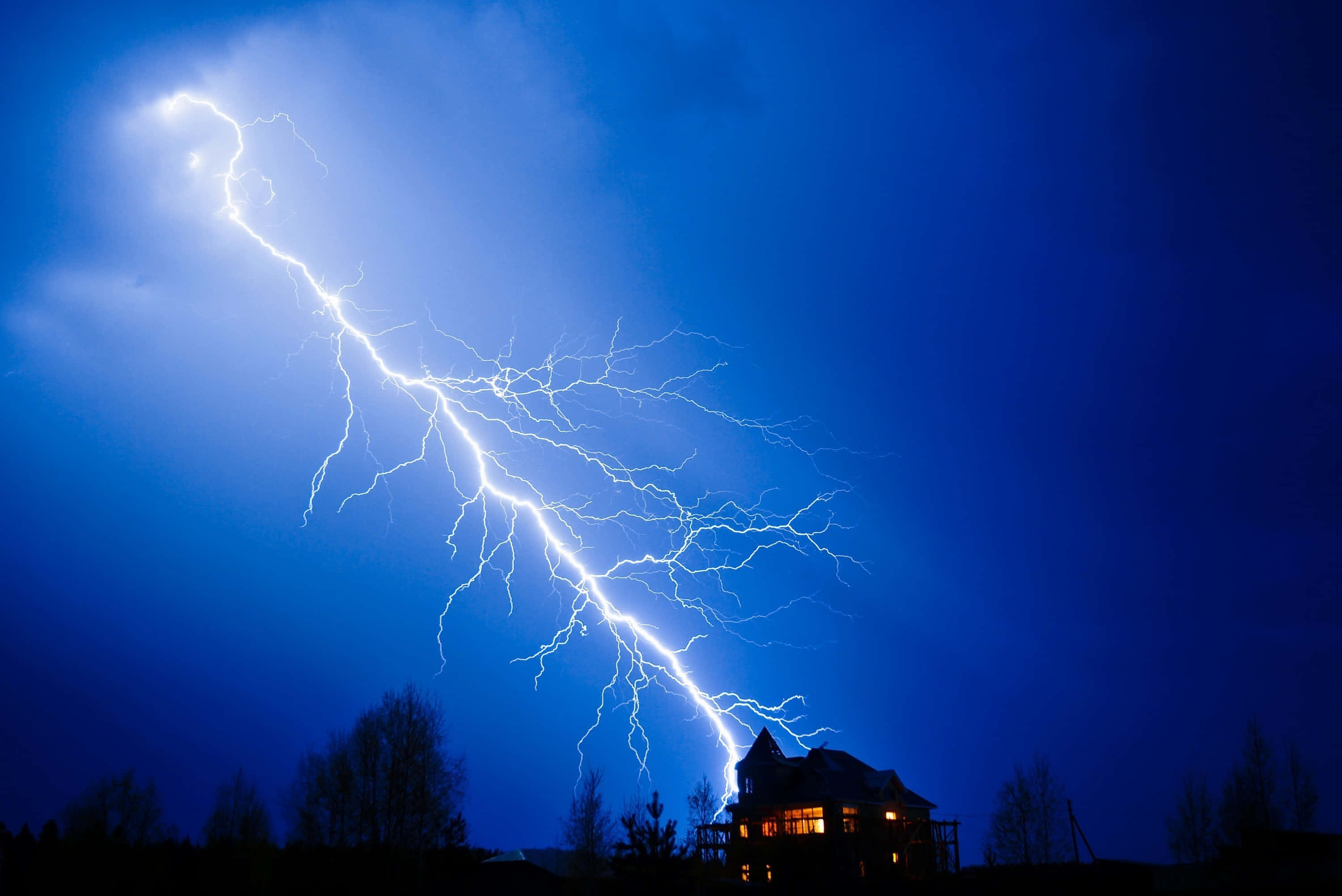 閃電 桌布 壁紙 — 一道明亮的閃電在深藍色的夜空中分叉，劃過一座在暴風雨中映襯出輪廓的溫暖燈光房屋。