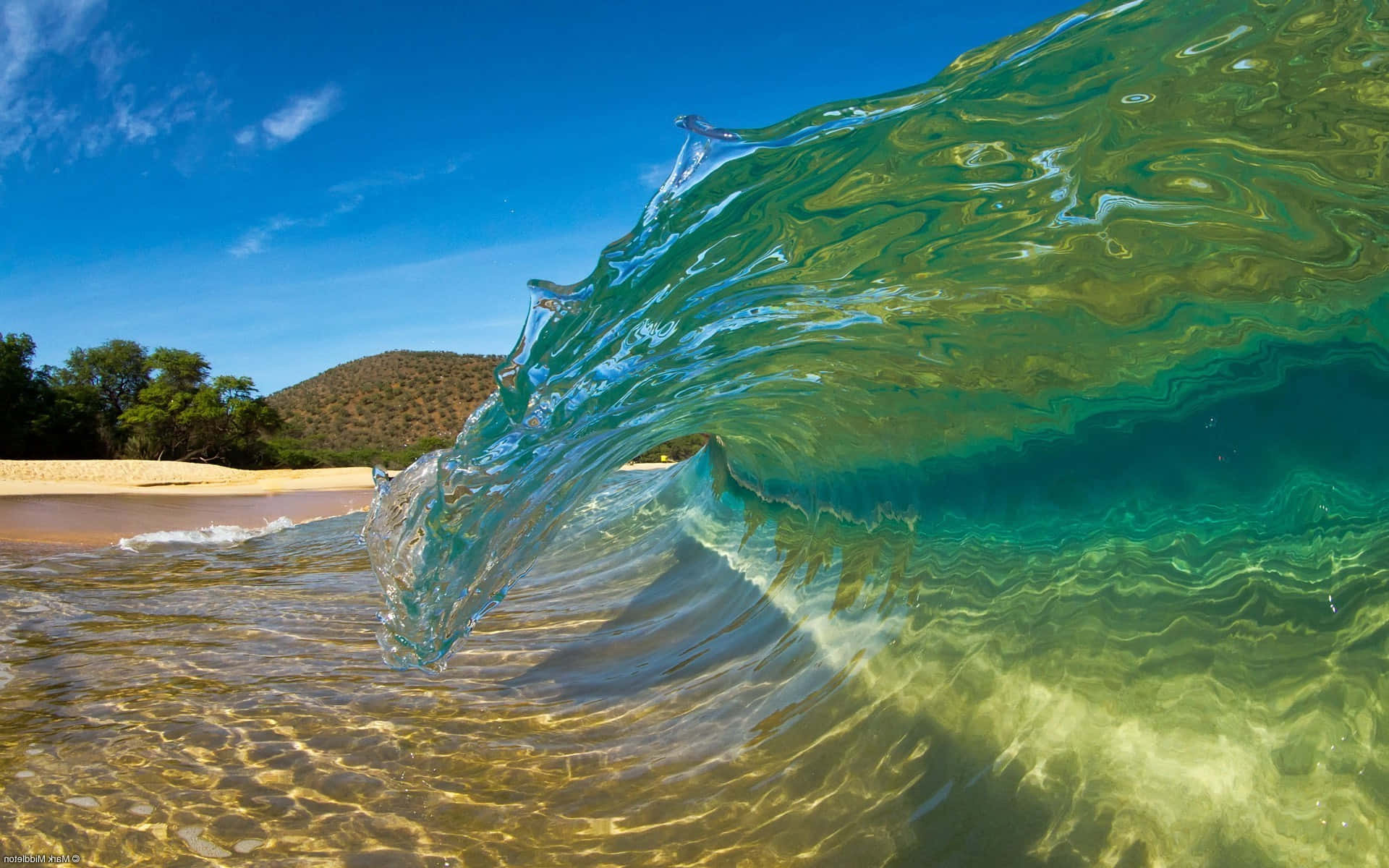 Beach Waves 桌布 壁紙 — 一道晶瑩剔透的碧綠色海浪在陽光普照的沙灘上翻捲，遠方有翠綠的山丘，天空湛藍明亮。