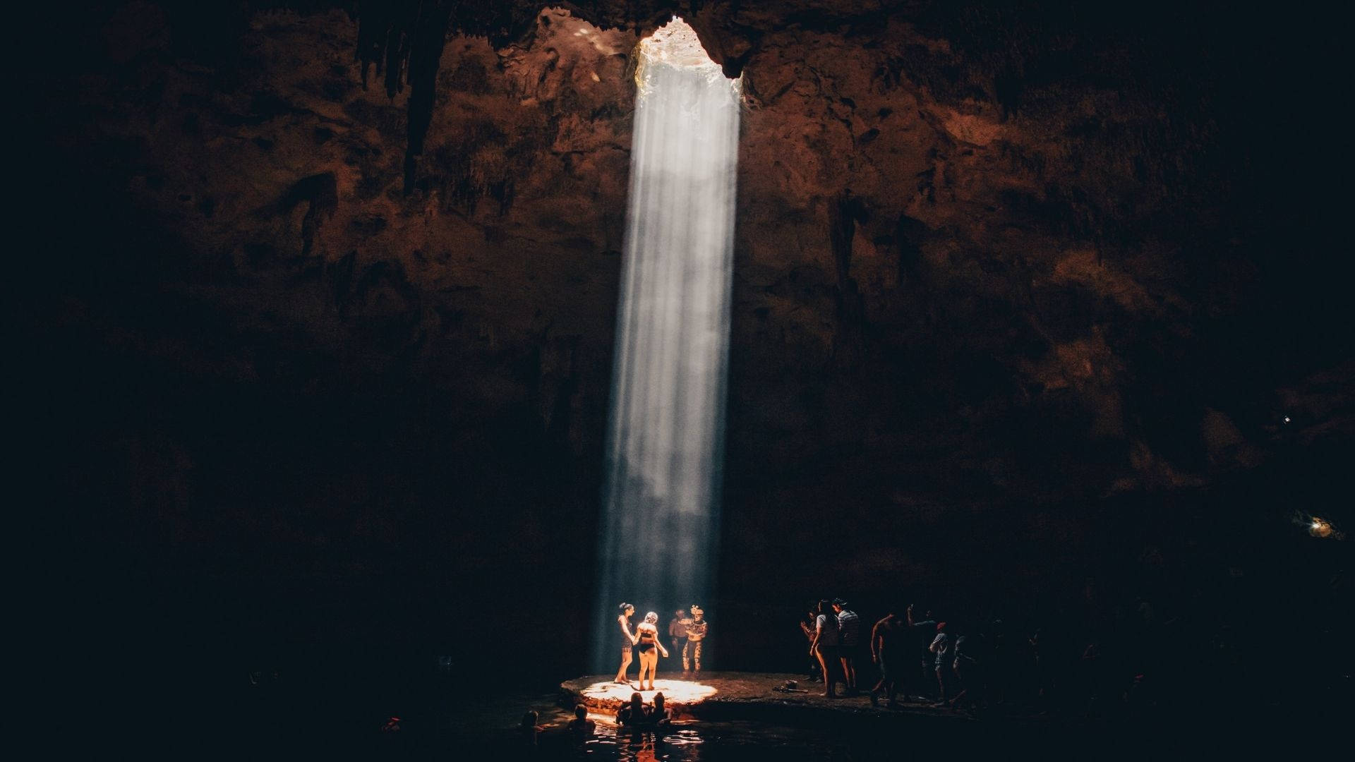 Cave 桌布 壁紙 — 一道狹窄的陽光穿透黑暗的洞穴天花板，照亮了瀑布和下方的人影。