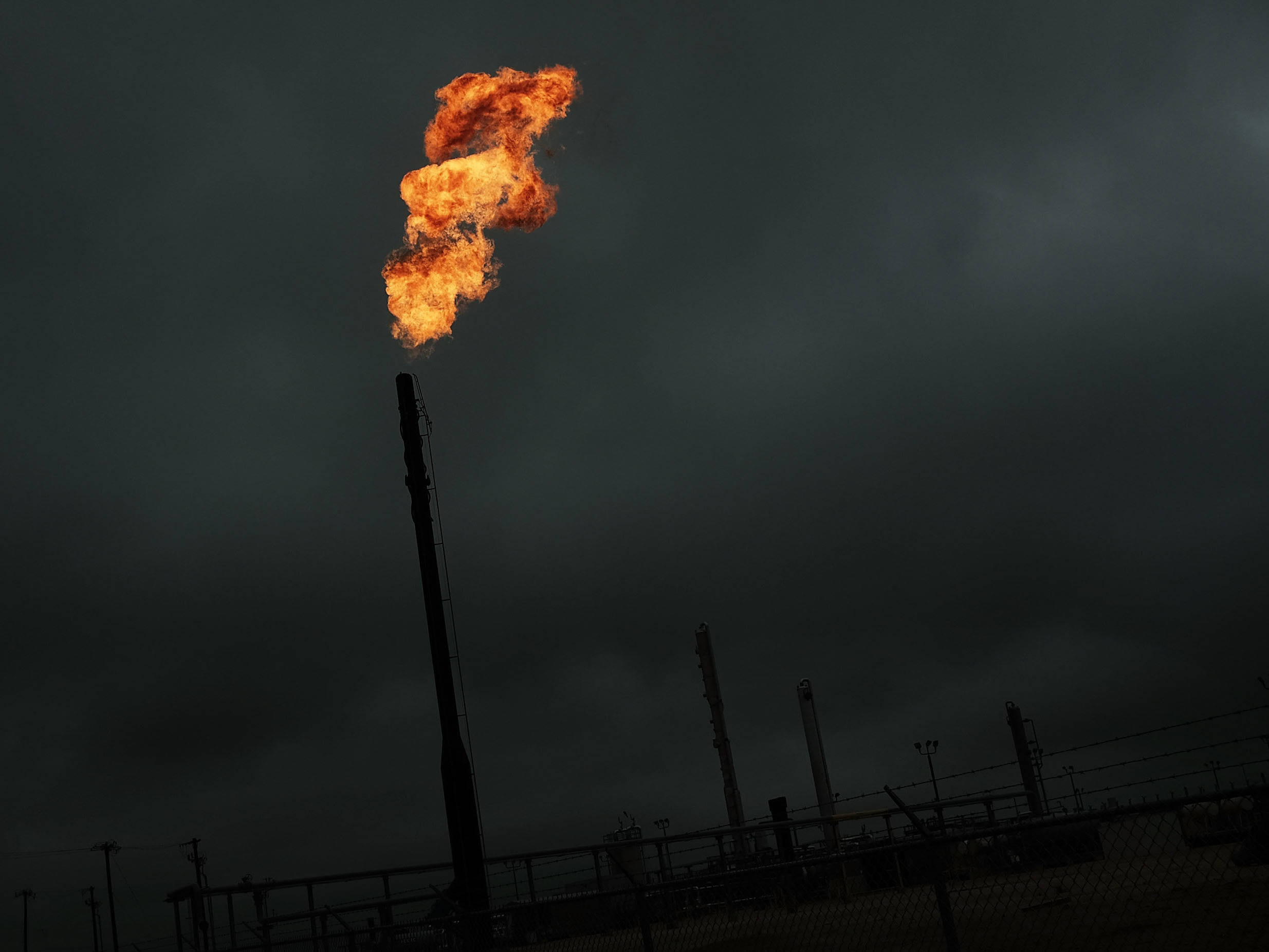 Gas 桌布 壁紙 — 一道猛烈的橙色火焰從高聳的工業煙囪噴出，背景是黑暗風暴的天空，下方是有圍欄的煉油廠綜合體。