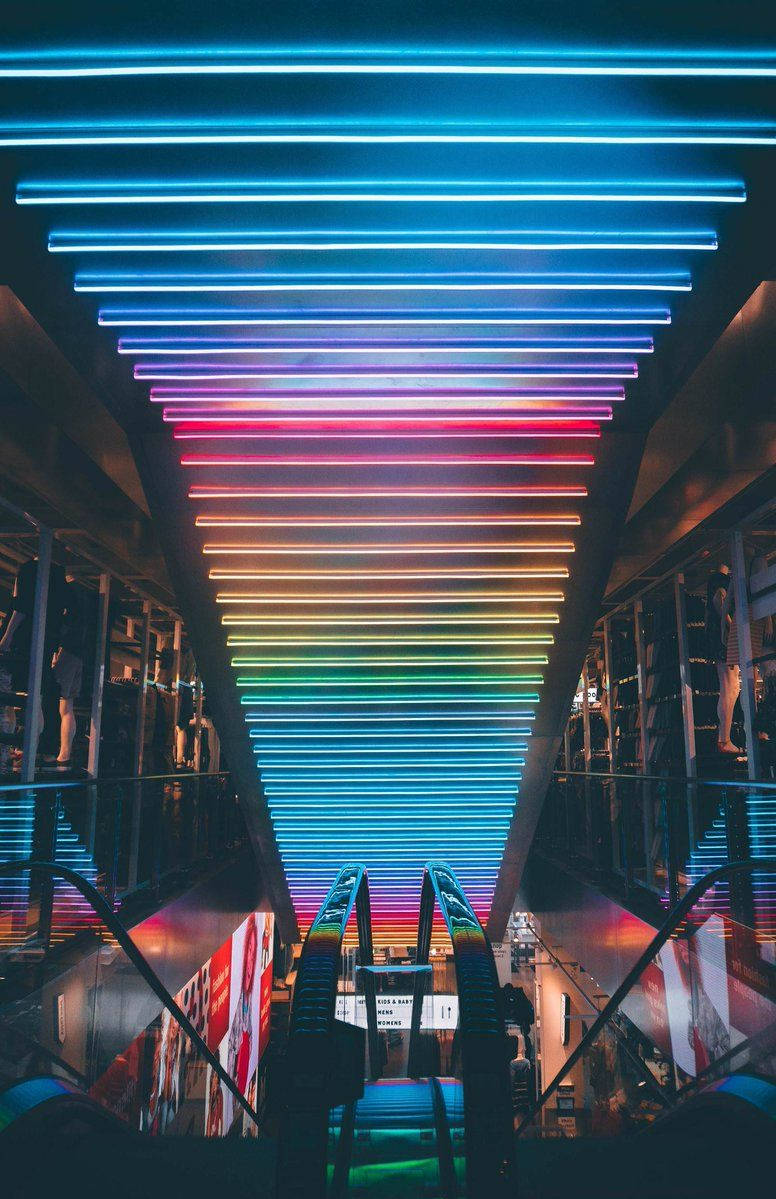 lights 桌布 壁紙 — 一道由霓虹燈組成的鮮豔彩虹，在現代城市交通空間的自動扶梯上方形成一個發光的三角形天花板。