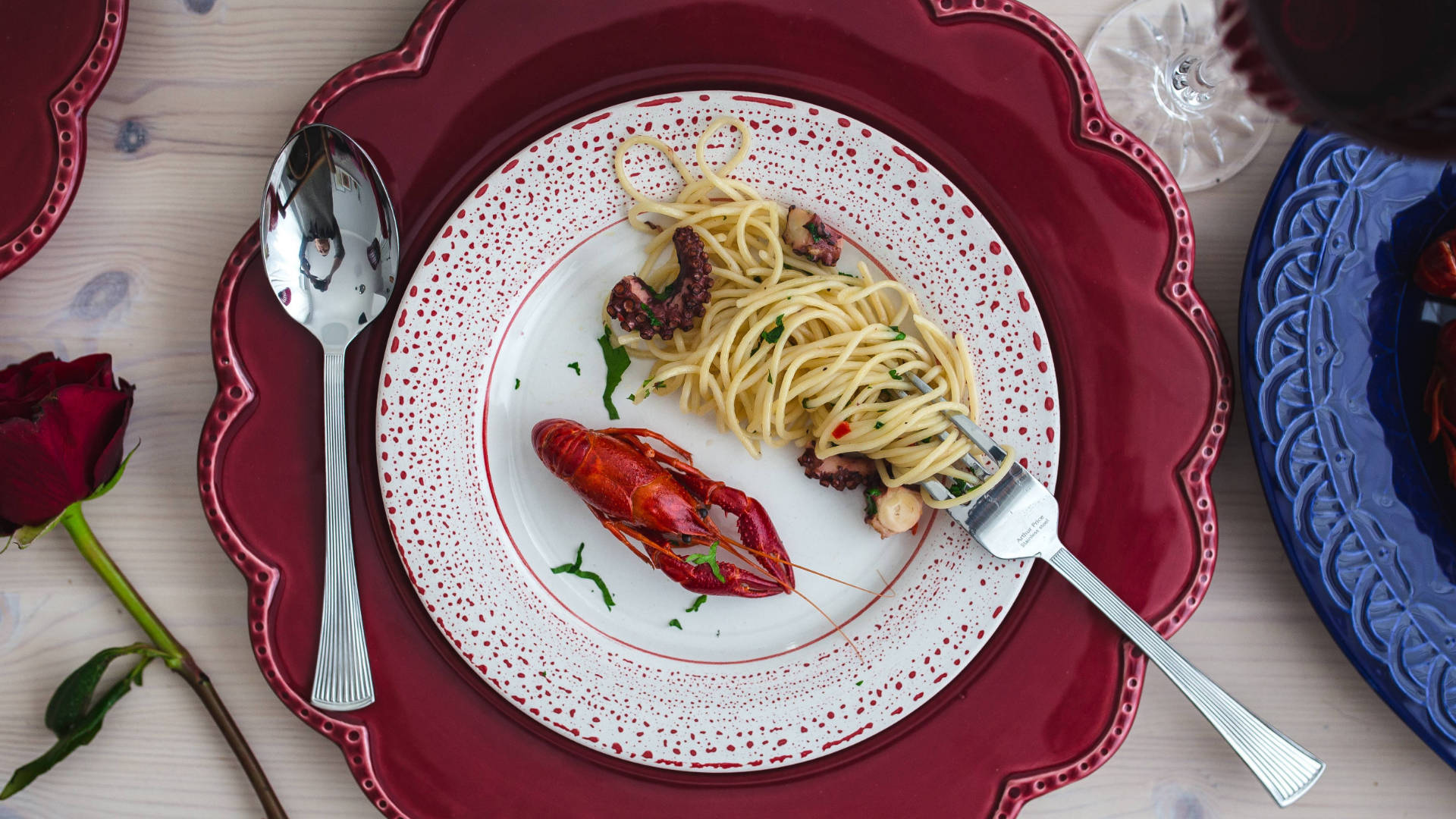 Pasta 桌布 壁紙 — 一道豪華的海鮮義大利麵菜餚，配有龍蝦和章魚，盛在紅色扇貝形盤子上，旁邊有玫瑰和酒杯。