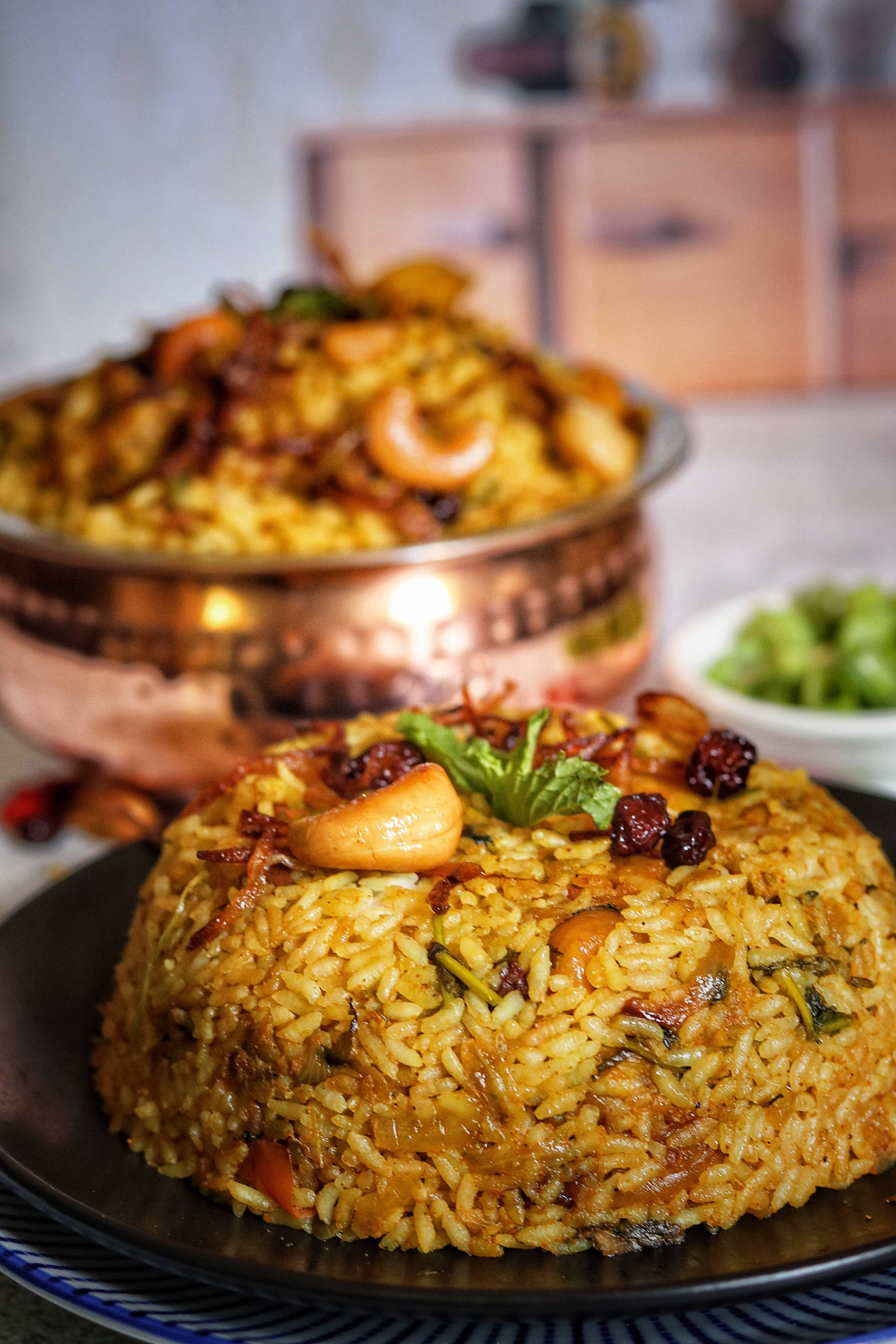 Delicious Food 桌布 壁紙 — 一道金色香料米飯，上面放有腰果、葡萄乾和薄荷，盛載於黑色碗中，背景有一個銅鍋。