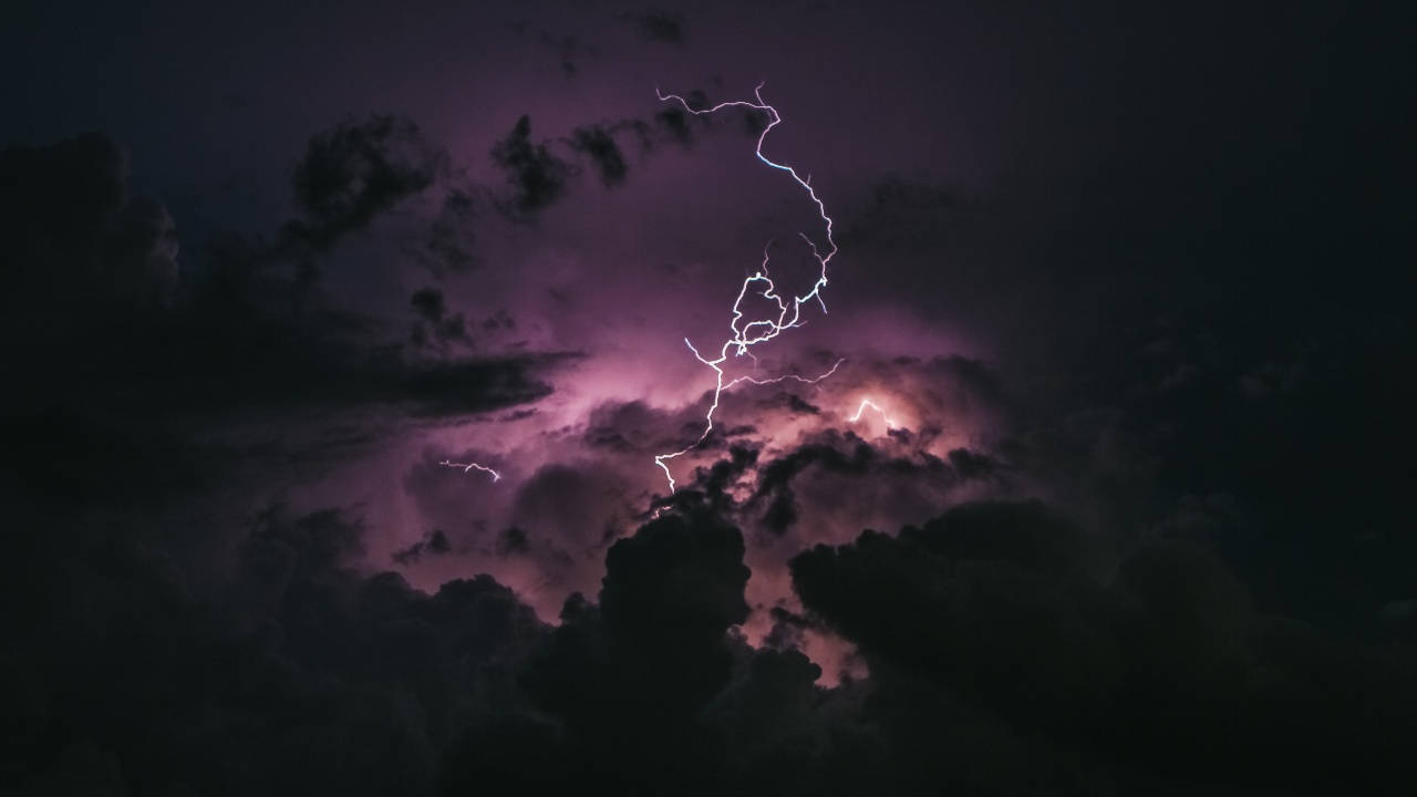 暗色電腦 桌布 壁紙 — 一道鋸齒狀的閃電在暴風雨的紫色天空中分叉，照亮了密集的雲層，形成一個戲劇性的夜晚場景。
