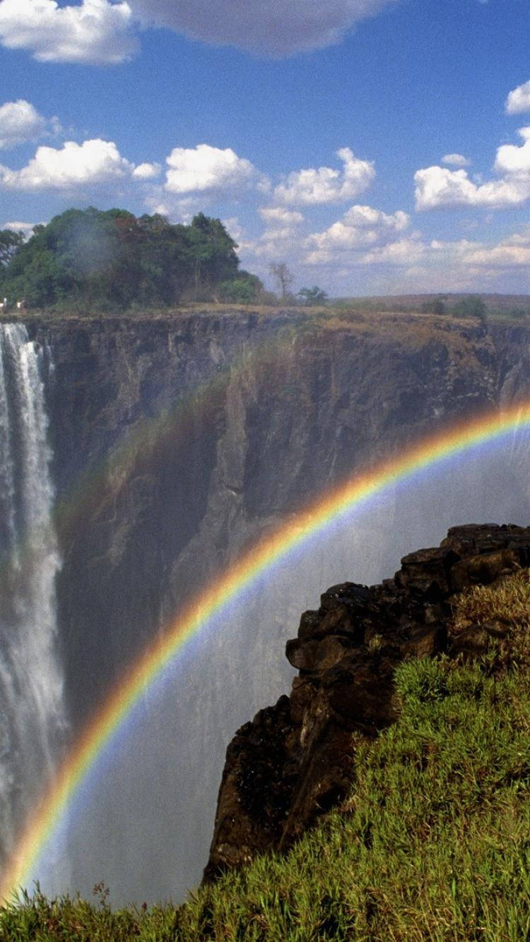 Zimbabwe 桌布 壁紙 — 一道雄偉的瀑布傾瀉入深峽谷，一道鮮豔的彩虹橫跨水霧，在藍天白雲下閃耀。