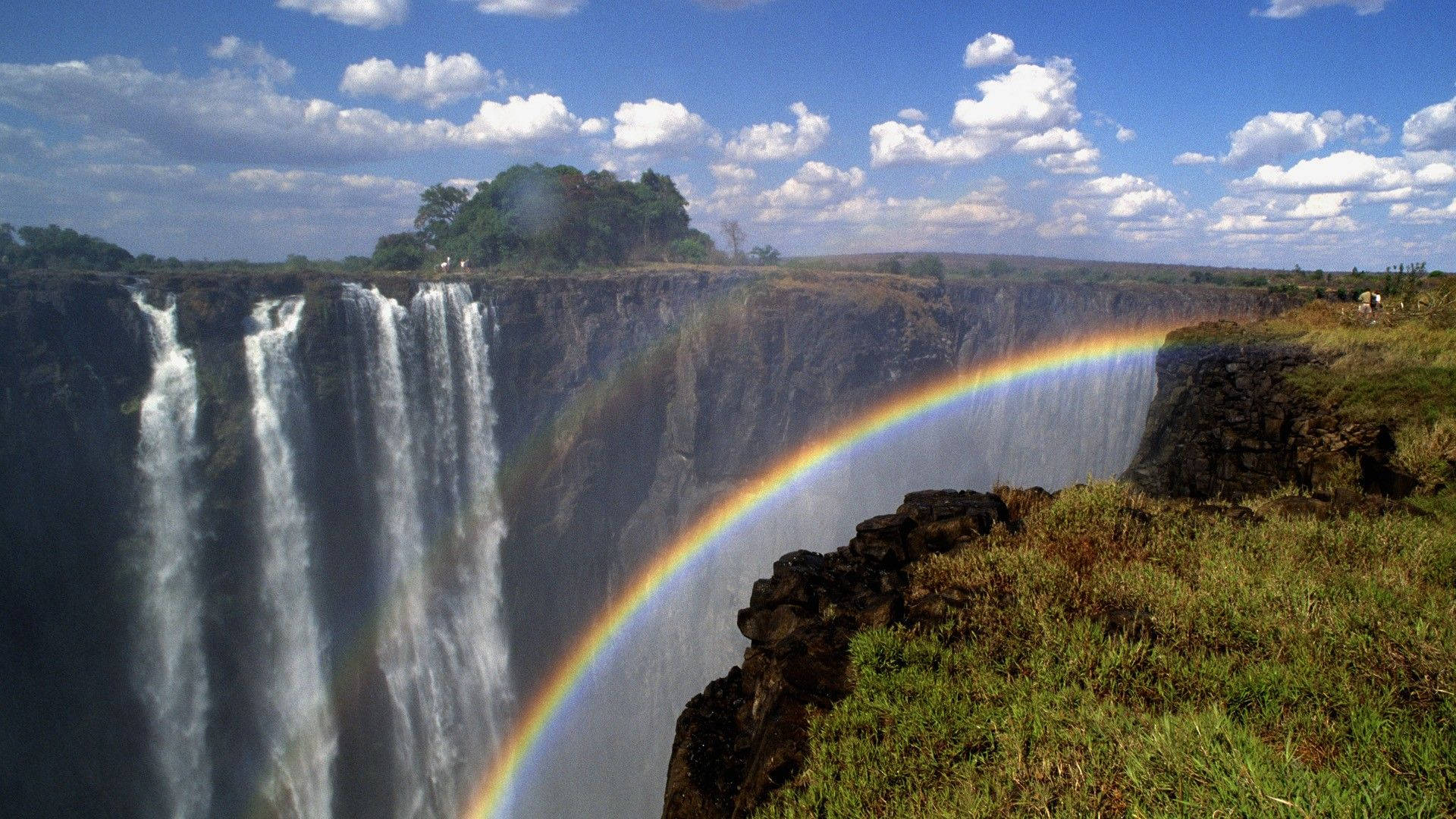 Zimbabwe 桌布 壁紙 — 一道雄偉的瀑布傾瀉入深峽谷，明亮的彩虹在霧氣中拱起，映襯著湛藍的天空。