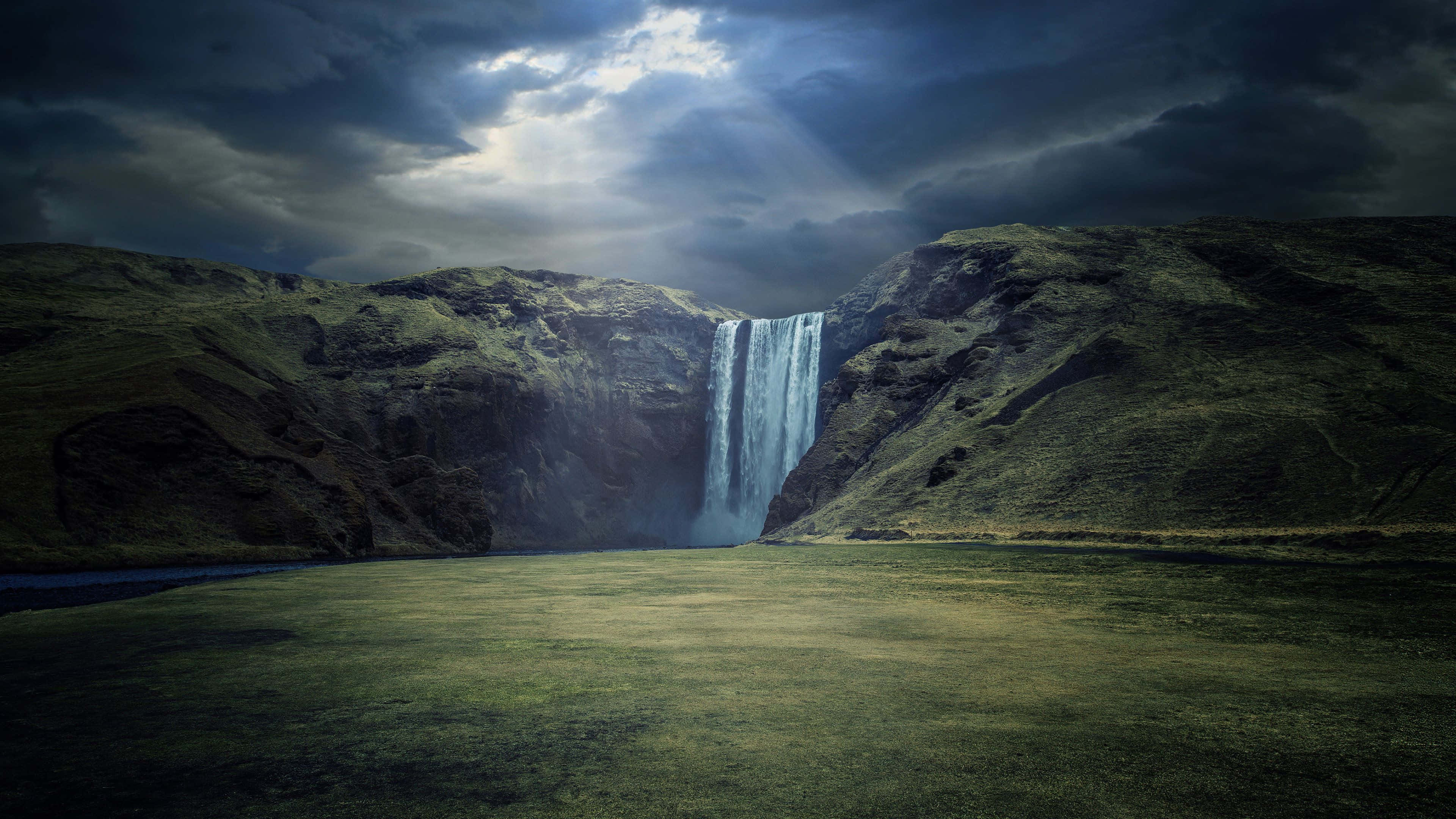4K Ultra HD Landscape 桌布 壁紙 — 一道雄偉的瀑布在崎嶇的綠色懸崖之間傾瀉而下，天空烏雲密佈，戲劇性十足，光線穿透雲層。