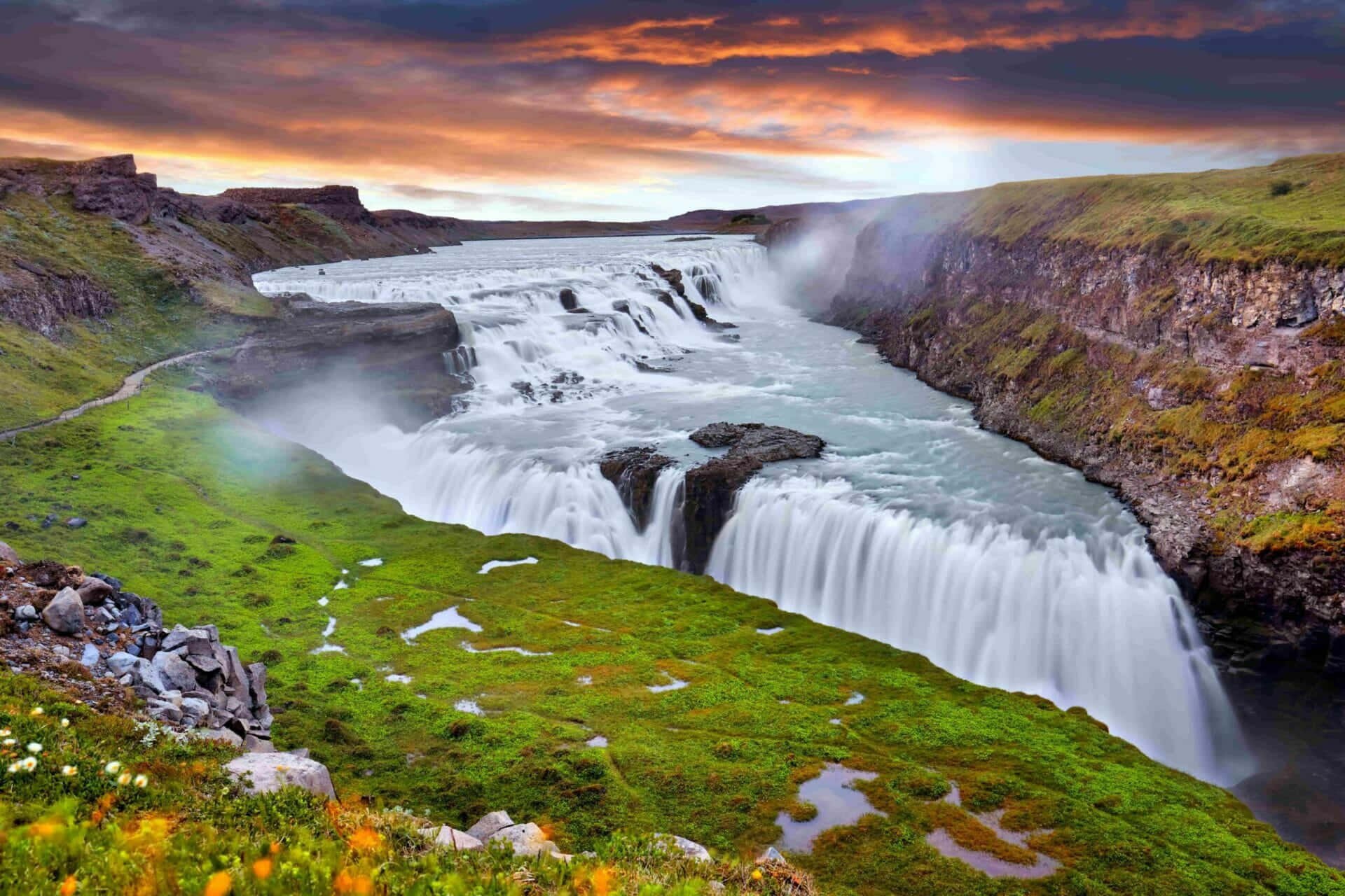 Iceland 桌布 壁紙 — 一道雄偉的瀑布在火紅的日落天空和戲劇性的雲彩下，流過鬱鬱蔥蔥的綠色峽谷。