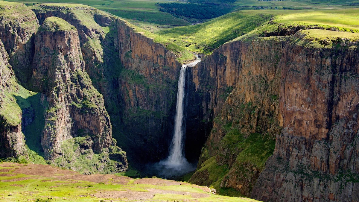 Lesotho 桌布 壁紙 — 一道雄偉的瀑布從崎嶇的懸崖傾瀉而下，落入被翠綠山丘環繞的深邃峽谷，在明亮的日光下格外壯觀。
