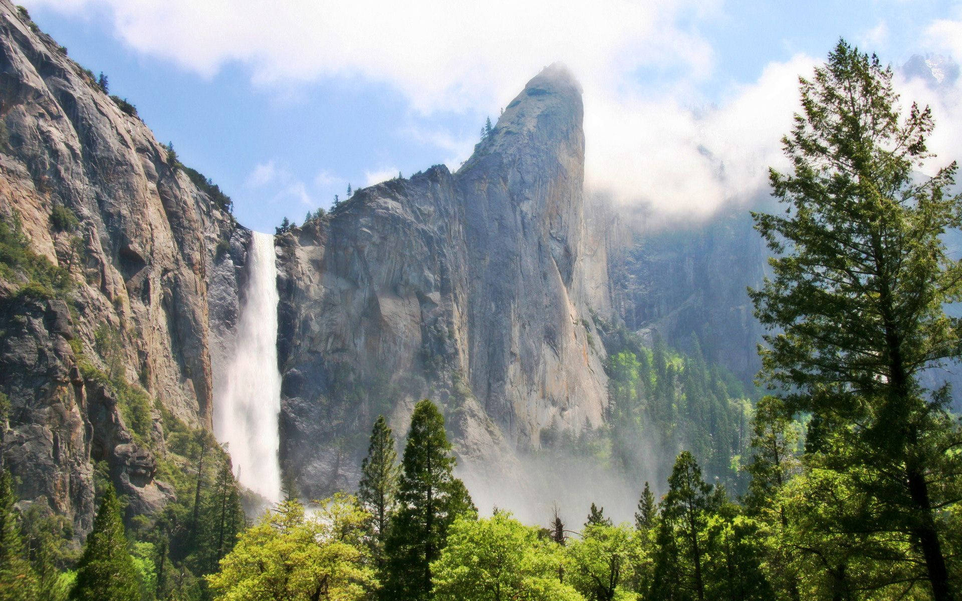 山景 桌布 壁紙 — 一道雄偉的瀑布從高聳的花崗岩懸崖傾瀉而下，周圍環繞著茂密的綠色松樹，頭頂是明亮的藍天和蓬鬆的白雲。