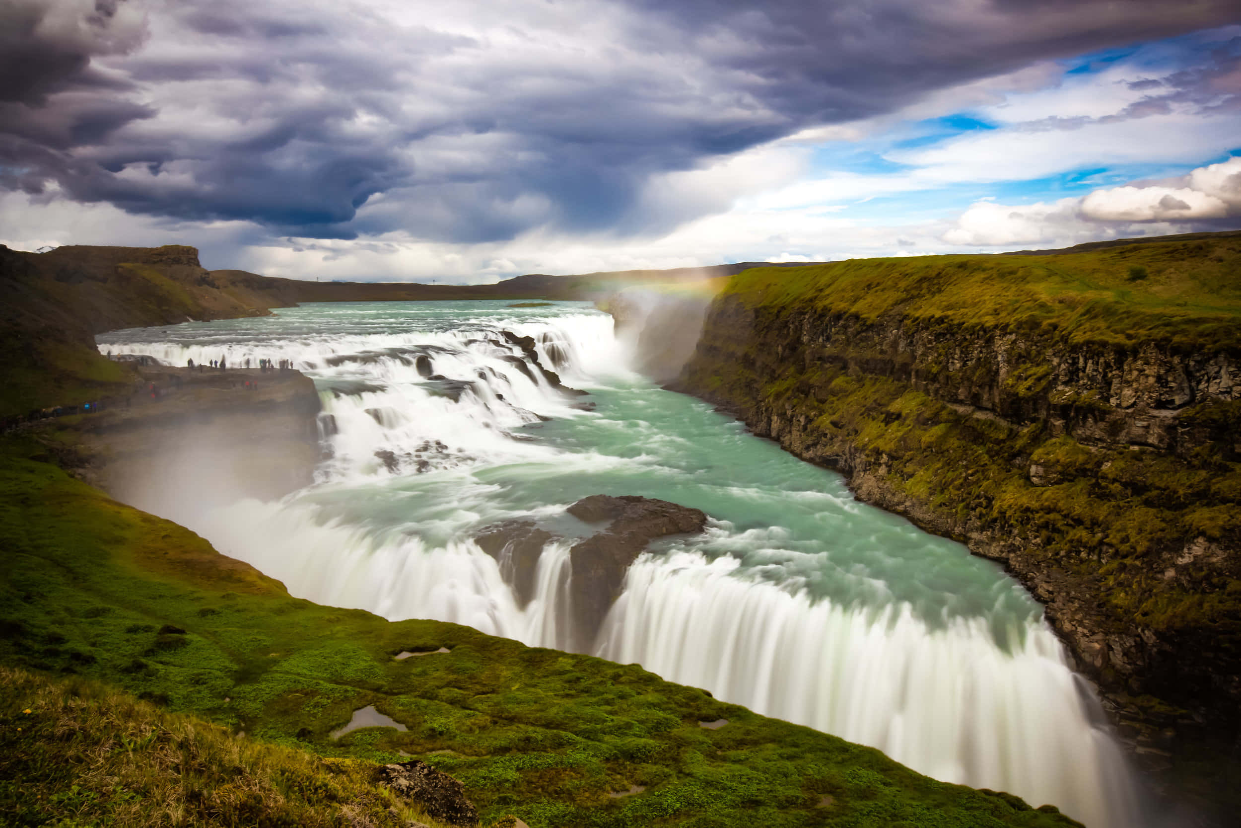 Iceland 桌布 壁紙 — 一道雄偉的瀑布流過長滿苔蘚的峽谷，在戲劇性的多雲天空下，一道淡淡的彩虹拱起在薄霧之上。