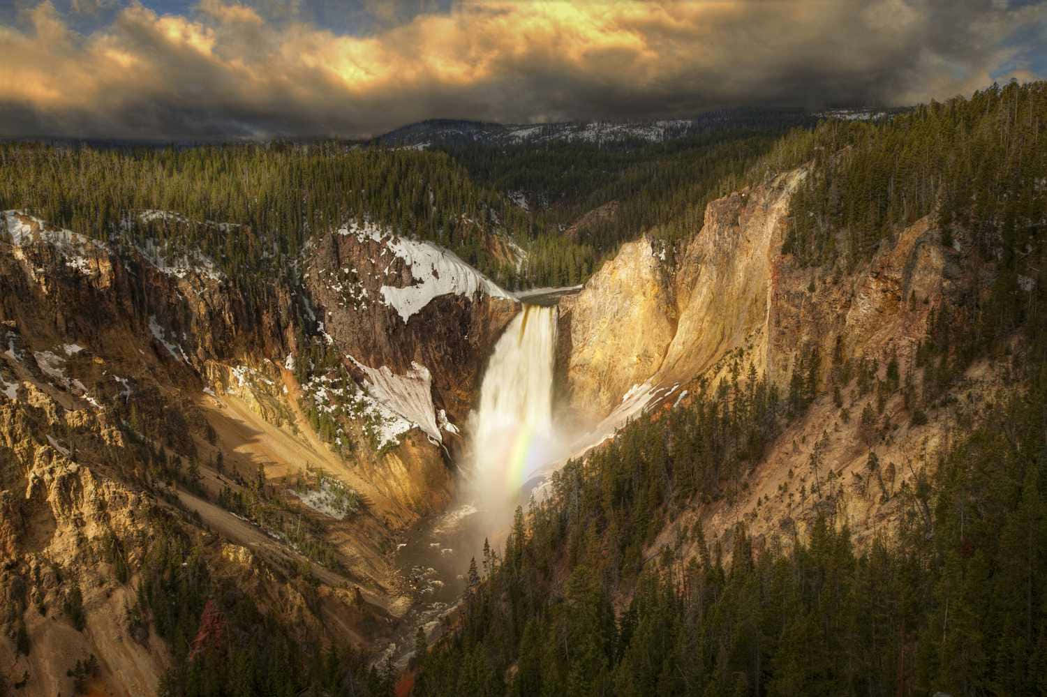 Yellowstone 桌布 壁紙 — 一道雄偉的瀑布穿過崎嶇的峽谷，兩旁是白雪覆蓋的懸崖和茂密的常綠森林，在戲劇性的金色日落天空下奔流而下。