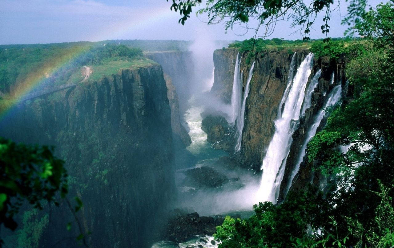 Zimbabwe 桌布 壁紙 — 一道雄偉的瀑布穿過深邃的峽谷，兩旁是茂密的綠植，一道鮮豔的彩虹橫跨在霧氣繚繞的懸崖之上。