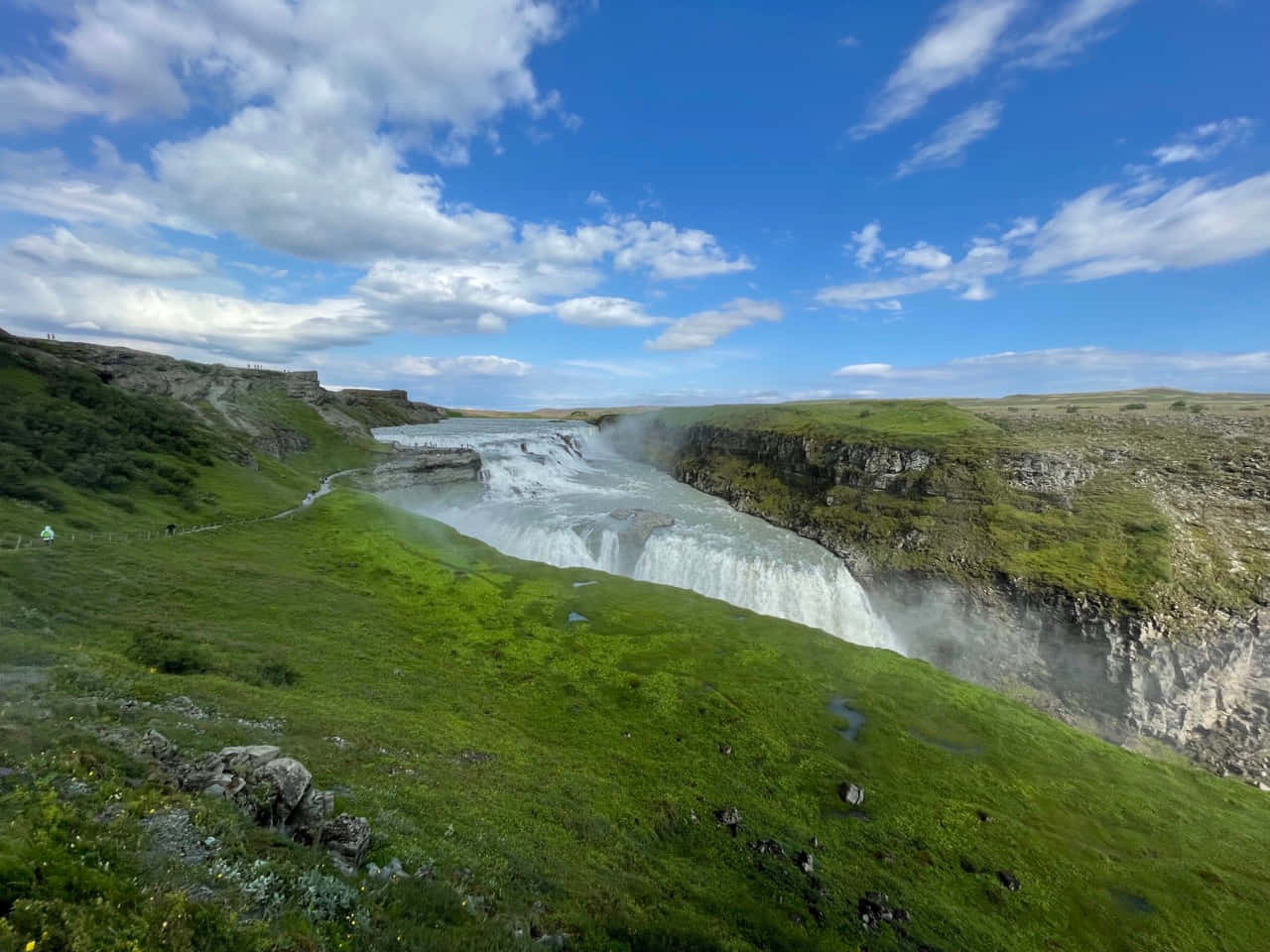 Iceland 桌布 壁紙 — 一道雄偉的瀑布穿過深邃的峽谷，周圍是鬱鬱蔥蔥的綠色山丘，在明亮的藍天下有蓬鬆的白雲。