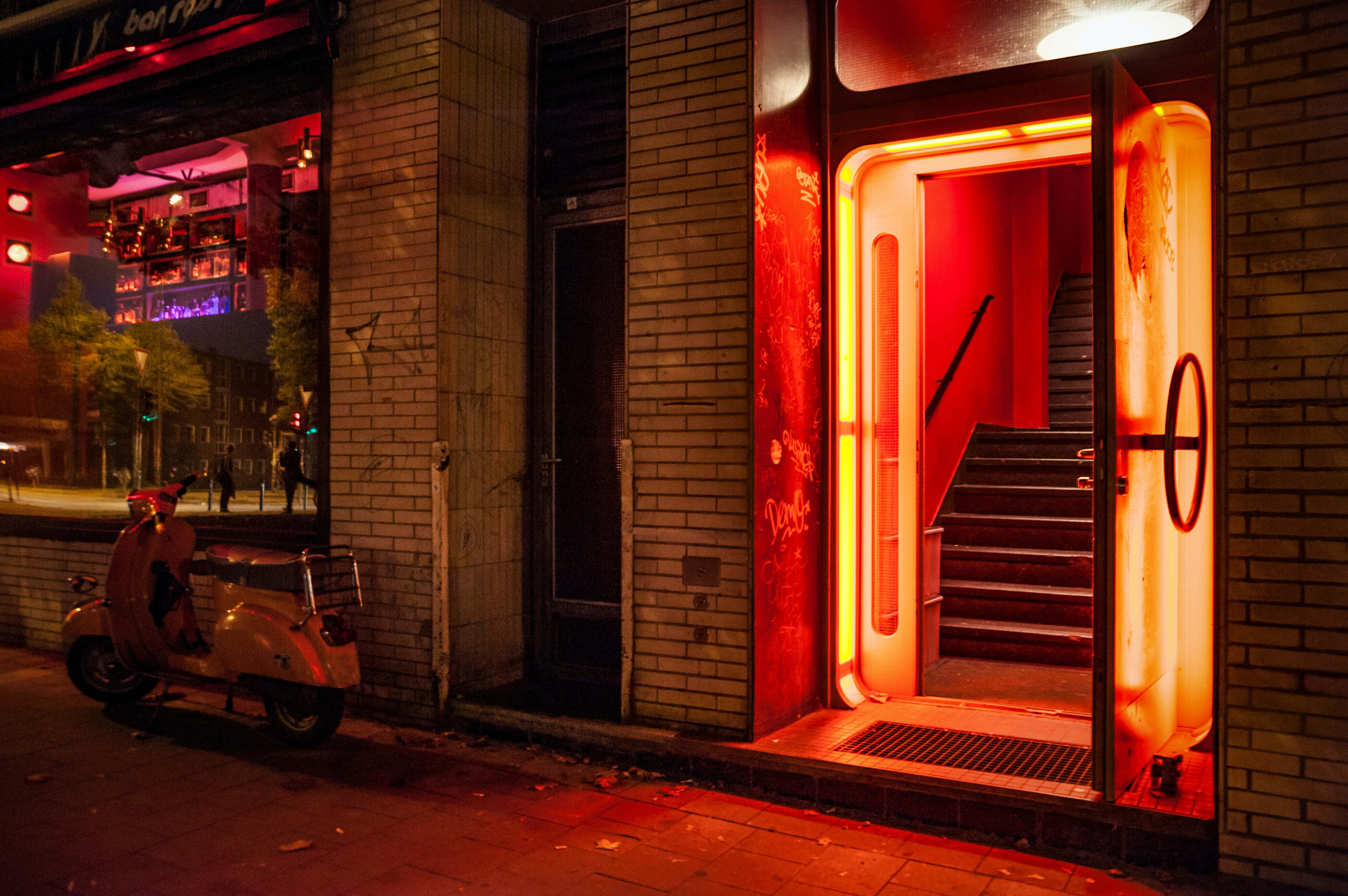 Nightclub 桌布 壁紙 — 一道霓虹燈照亮的門口，沐浴在紅色光芒中，顯露出內部的樓梯，而一輛復古機車則停在夜晚的城市街道上。