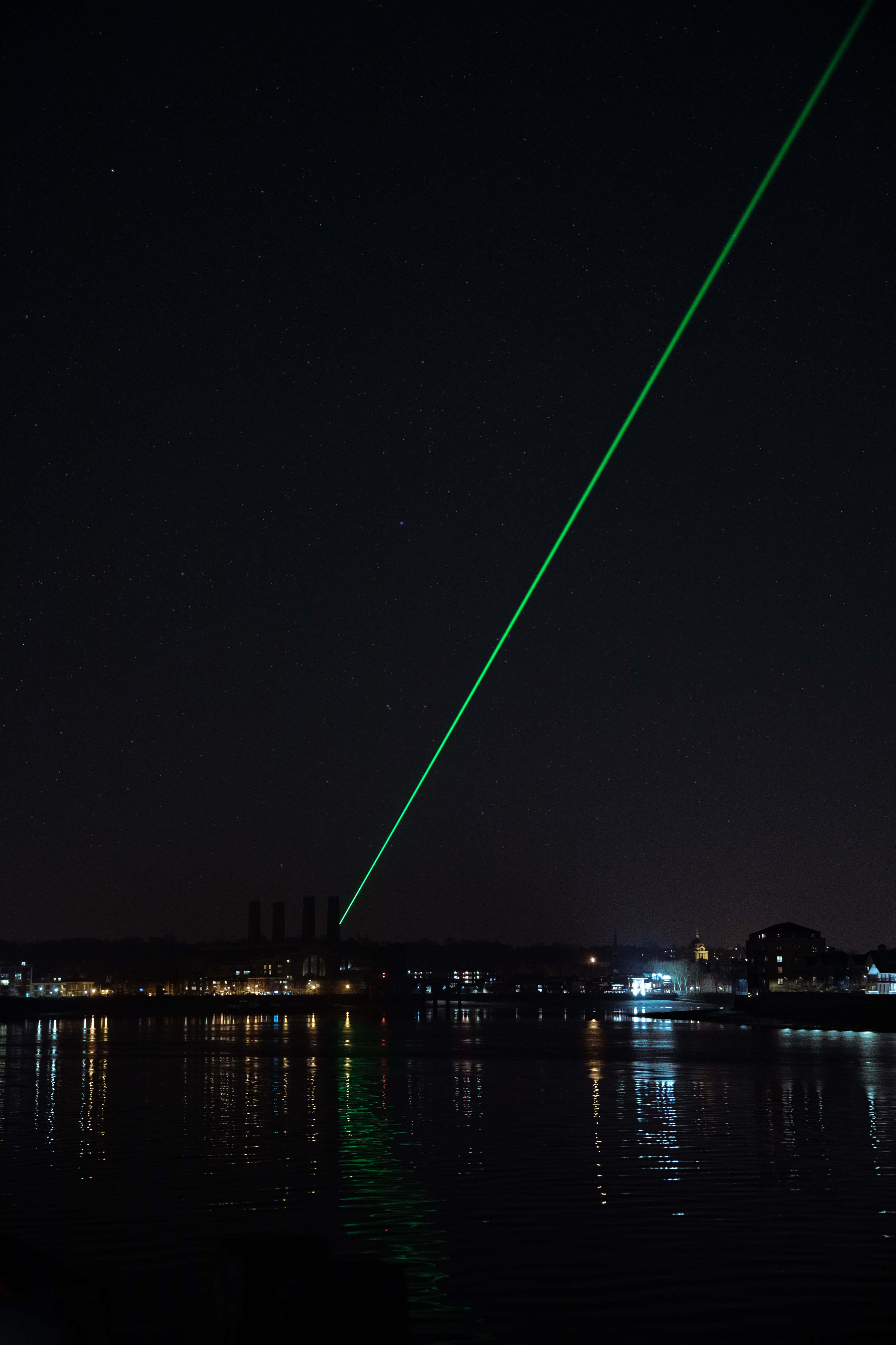 Laser 桌布 壁紙 — 一道鮮綠色雷射光束劃過繁星點點的夜空，下方平靜的水道倒映著城市燈光和工業建築。