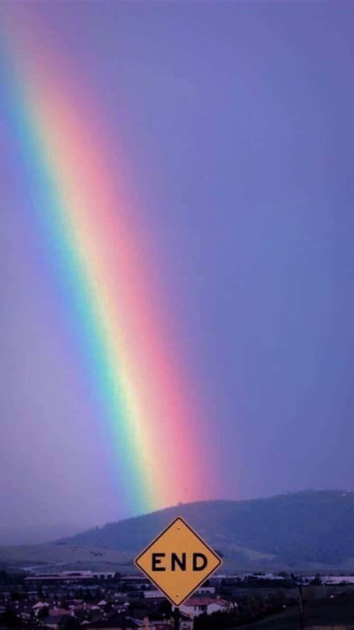 Aesthetic Rainbow Mobile 桌布 壁紙 — 一道鮮豔的彩虹拱起在小鎮上空，前景有一個黃色的「END」路牌，背景是紫色的暮色天空。