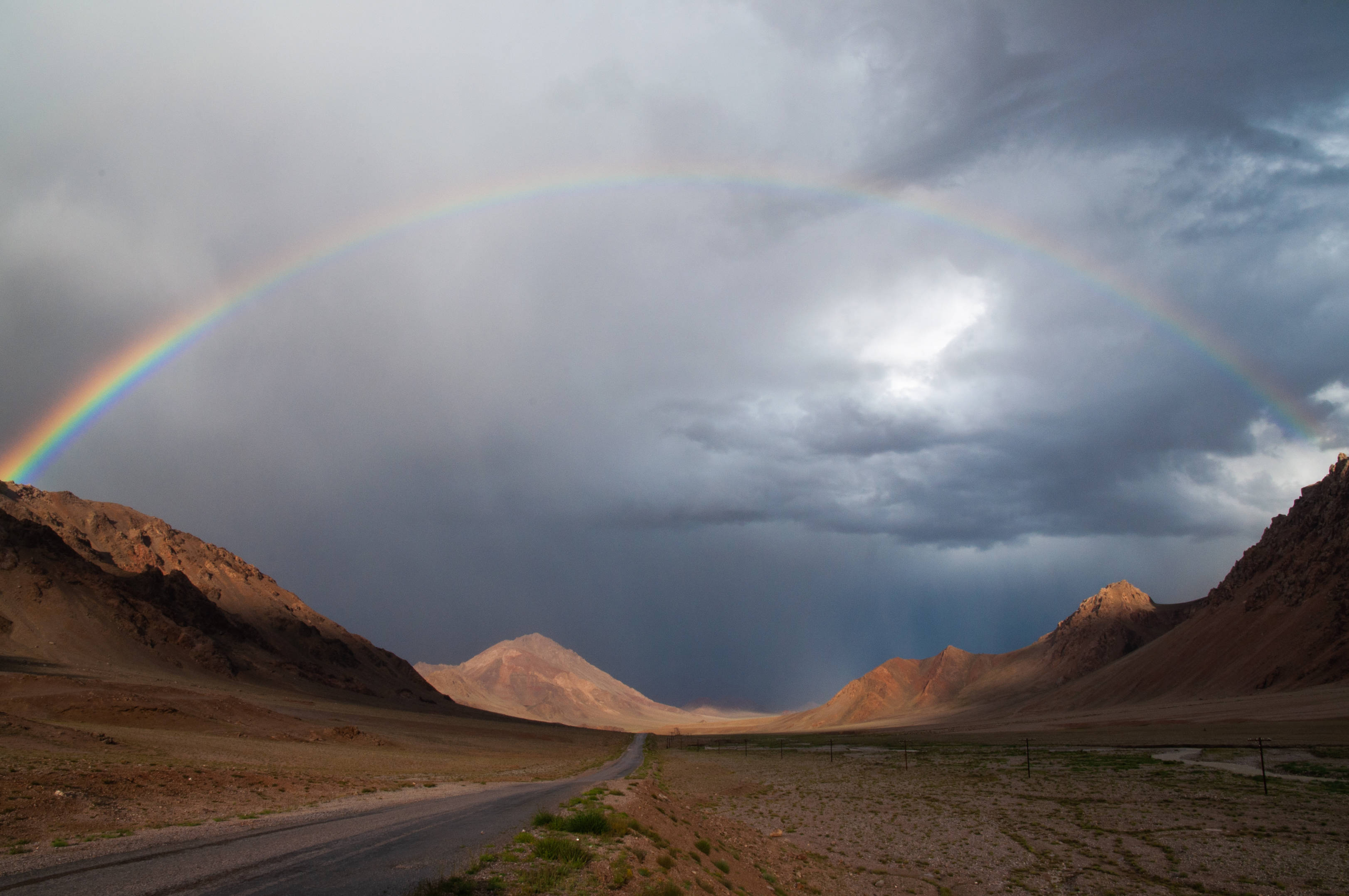 Tajikistan 桌布 壁紙 — 一道鮮豔的彩虹橫跨荒涼的山谷，雨在暴風雨的天空下落下。