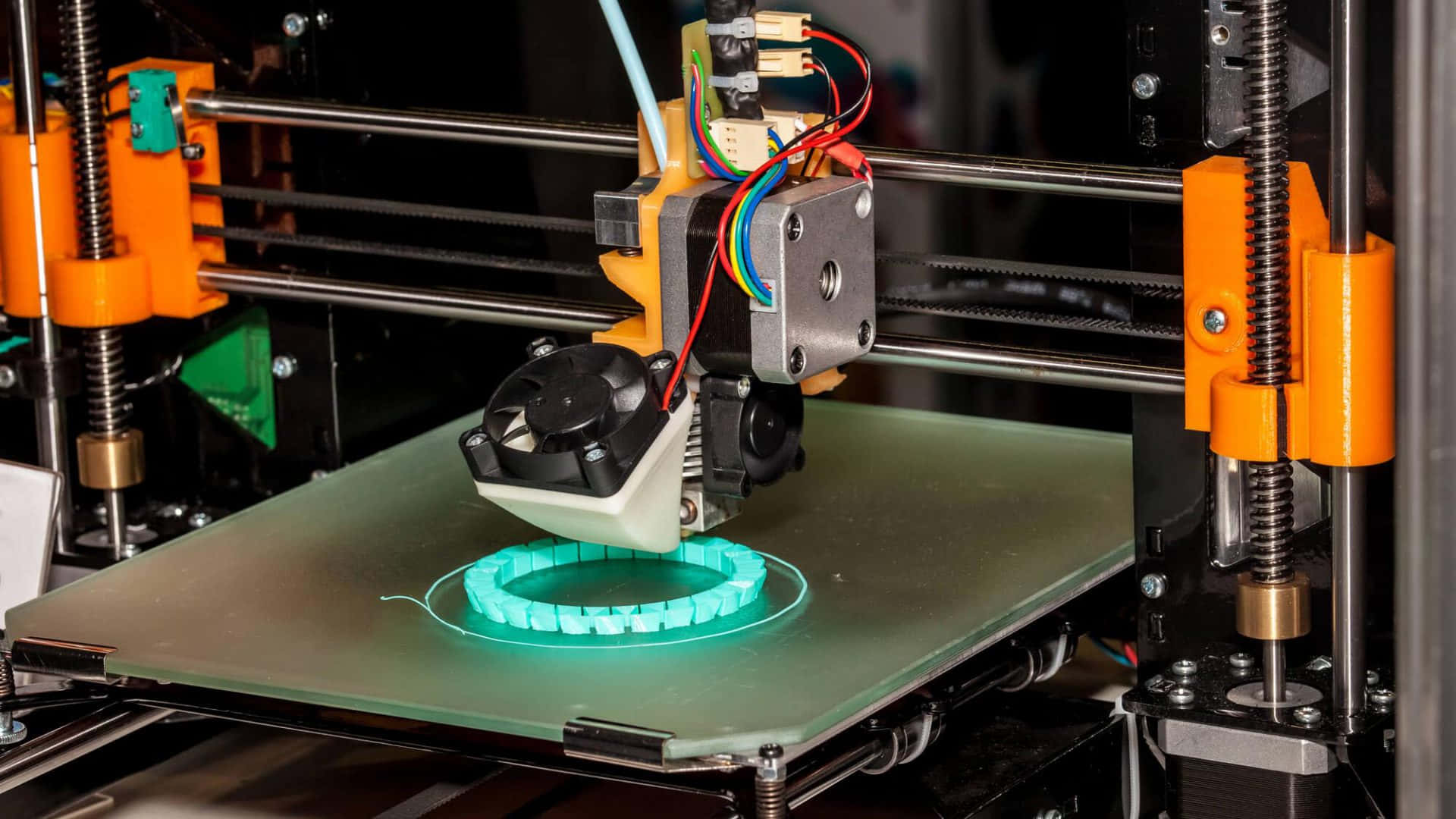 3d Printer 桌布 壁紙 — 一部3D列印機擠出發光的青綠色線材，在玻璃床上用橙色機械臂形成一個圓形齒輪。