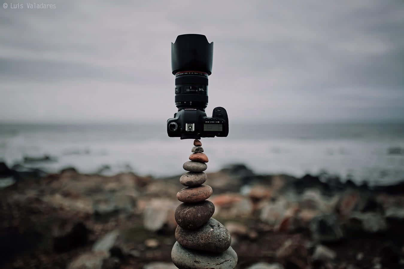 Dslr 桌布 壁紙 — 一部DSLR相機連鏡頭平衡在霧氣籠罩的海岸上一個不穩定的光滑石堆上。