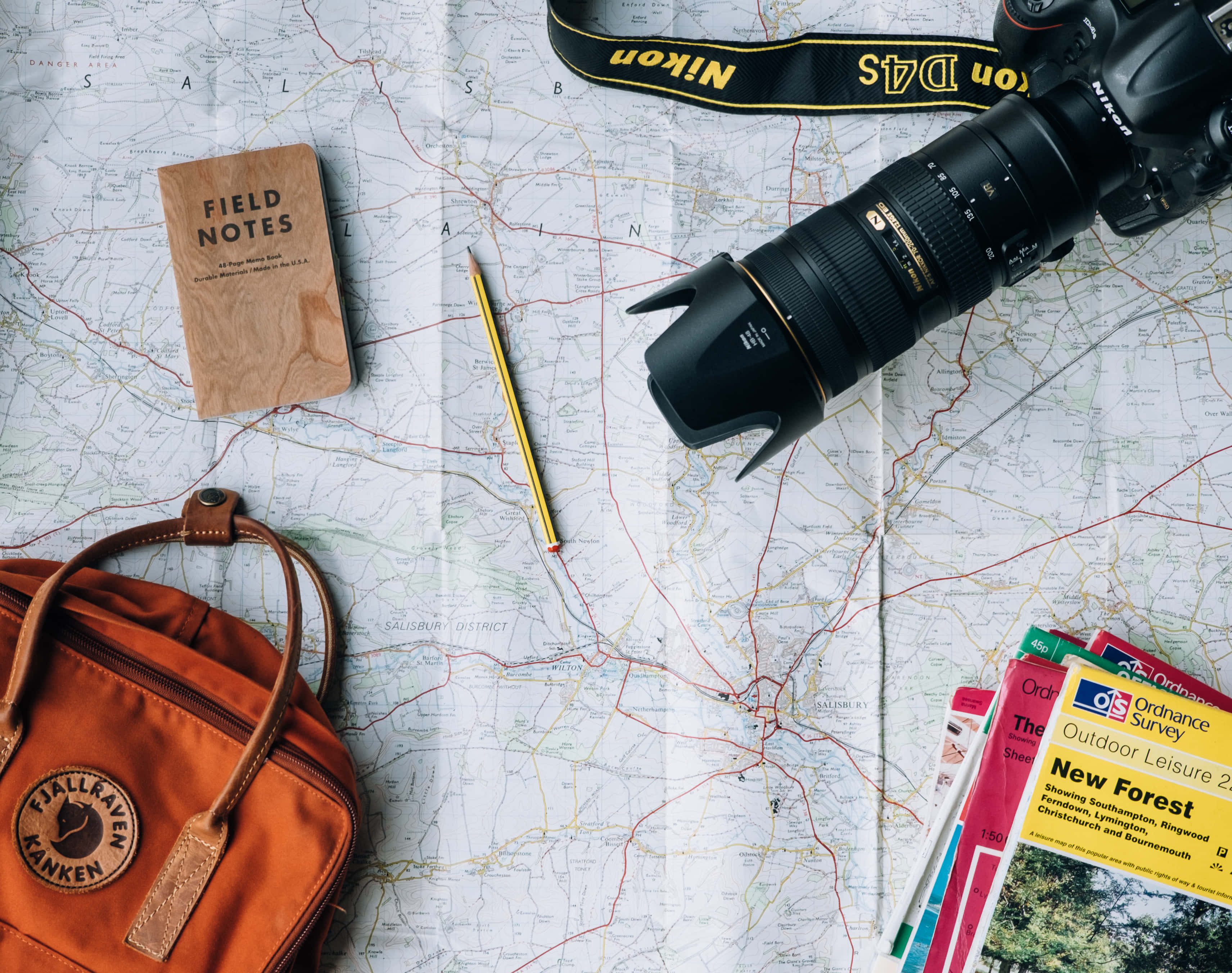 World Travel 桌布 壁紙 — 一部Nikon DSLR相機和一個橙色背包放在詳細的地形圖上，周圍有實地筆記和旅遊指南。