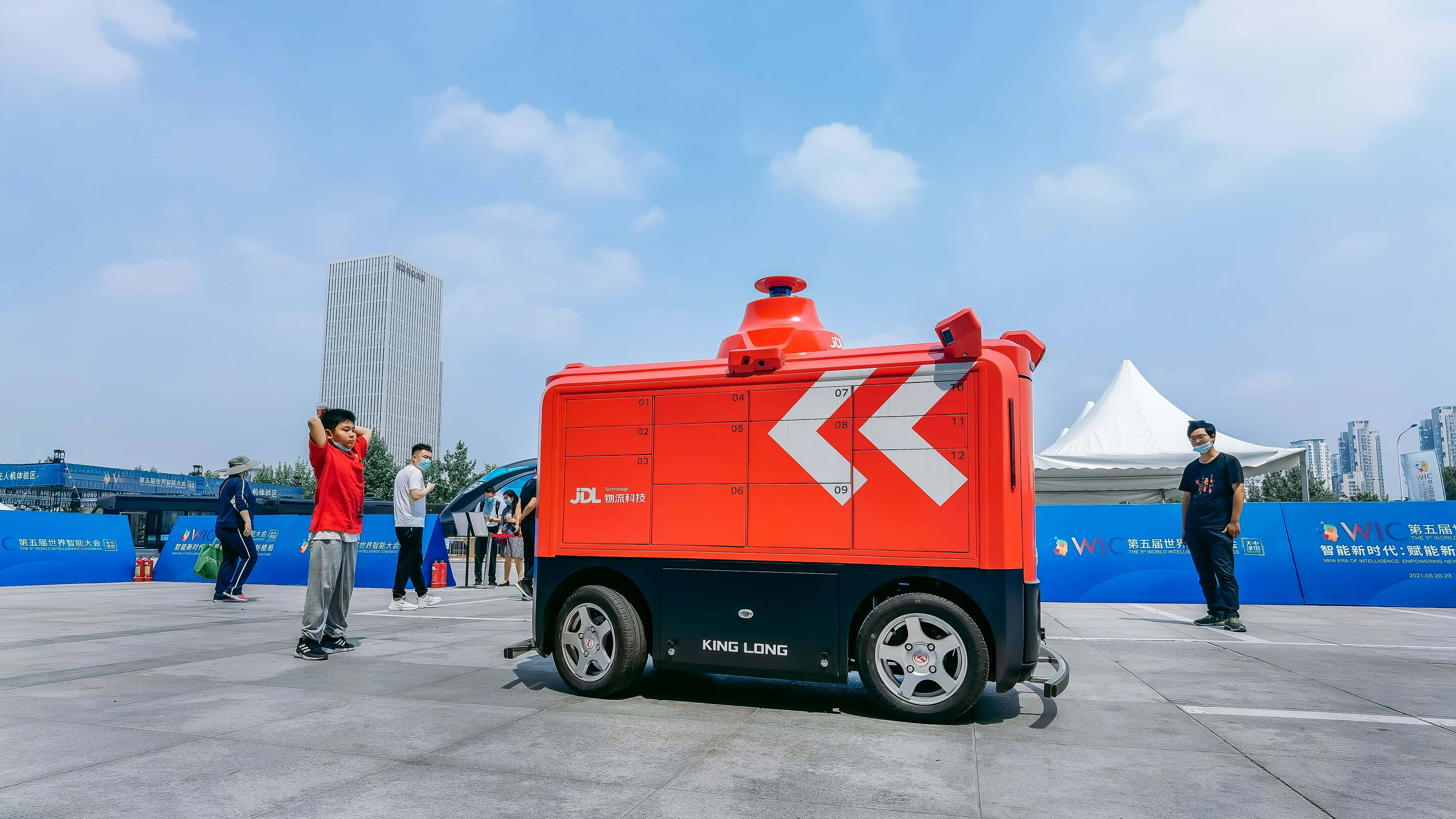 Truck 桌布 壁紙 — 一部亮橙色的自動送貨機器人帶有 'JDL' 和 'KING LONG' 品牌標誌，站在鋪砌的廣場上，在晴朗的藍天下，人們在附近觀看。