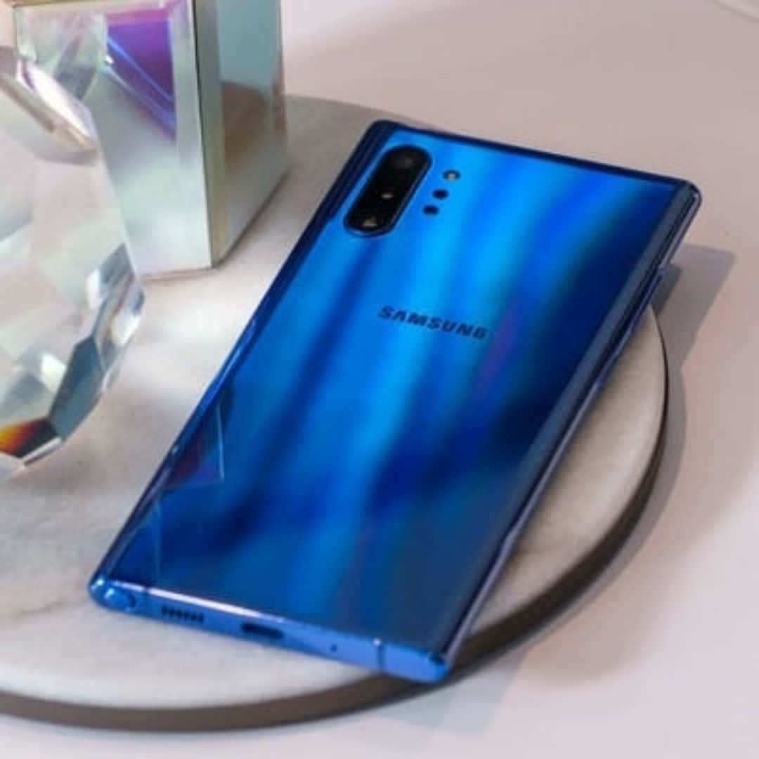 Note 10 桌布 壁紙 — 一部亮藍色Samsung智慧型手機放在大理石杯墊上，旁邊有一個多面玻璃裝飾品，在明亮燈光下。
